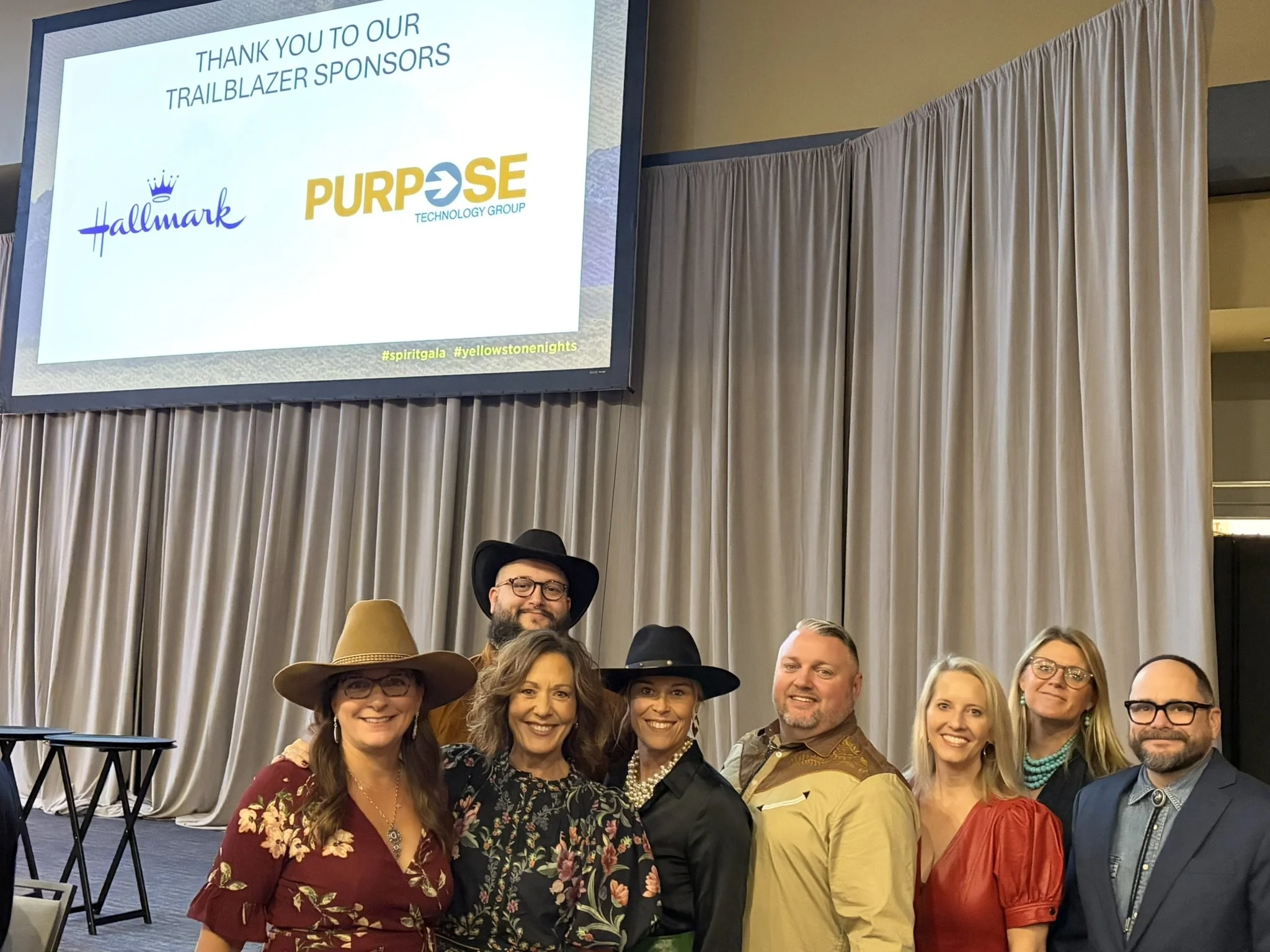 A group of people, mostly women, smiling and posing for a photo in front of a beige curtain at an event. On the screen behind them, there is a message thanking sponsors, including Hallmark and Purpose Technology Group, with hashtags #spiritgala and #