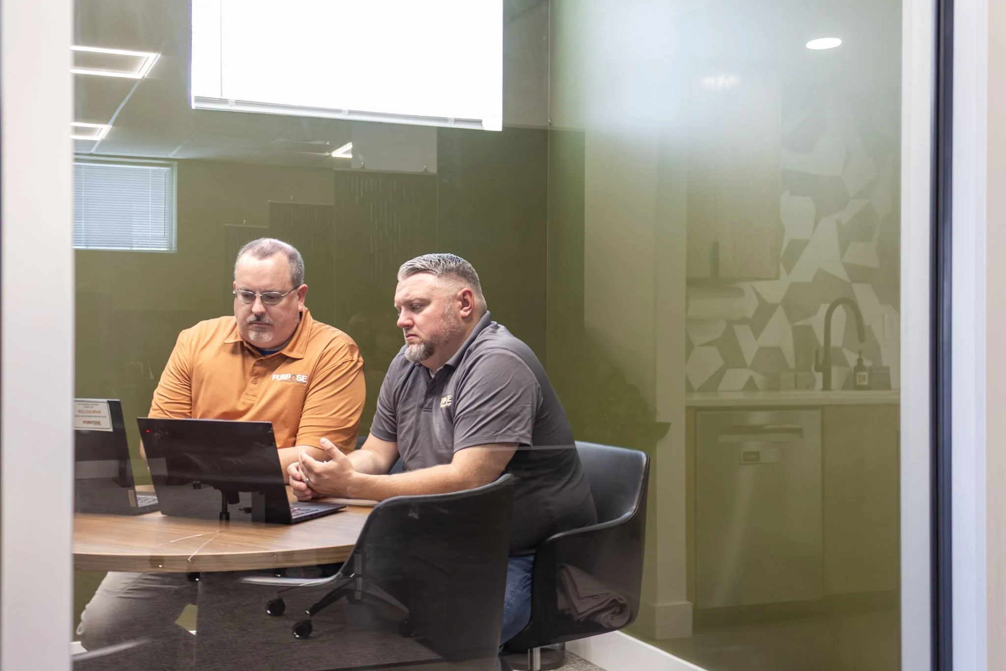 Two men sitting at a conference table looking at a laptop computer, seen through a glass wall in an office.