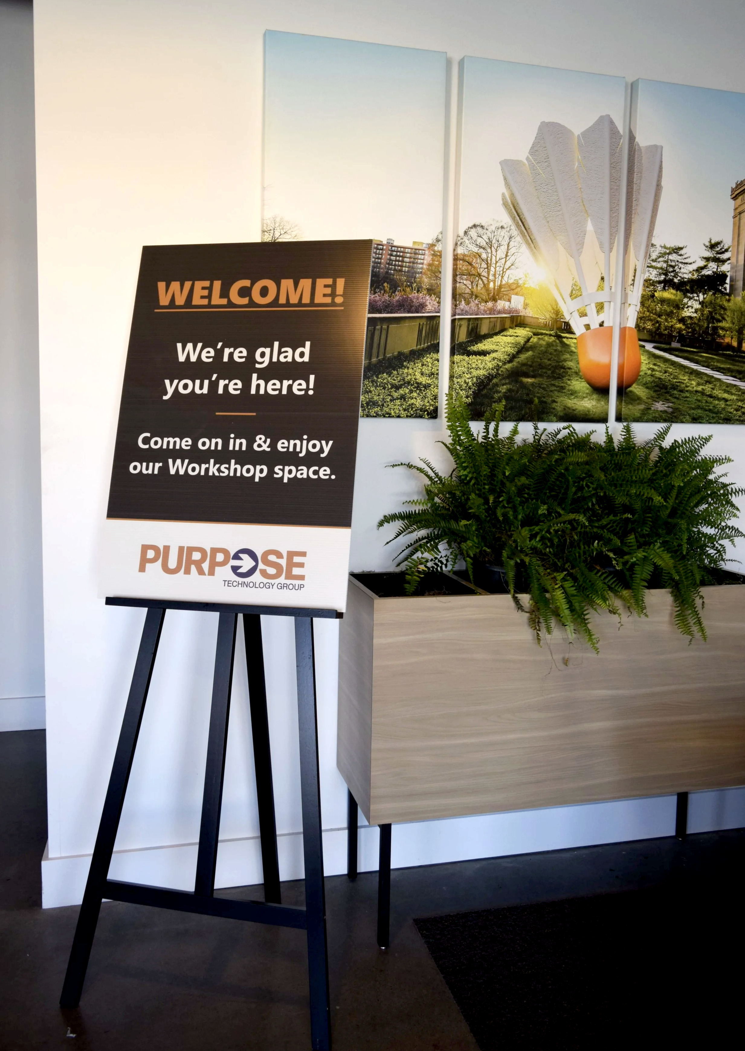 Welcome sign that says We're glad you're here! on easel in front of planter and photograph from the Nelson Atkins Museum in Kansas City.