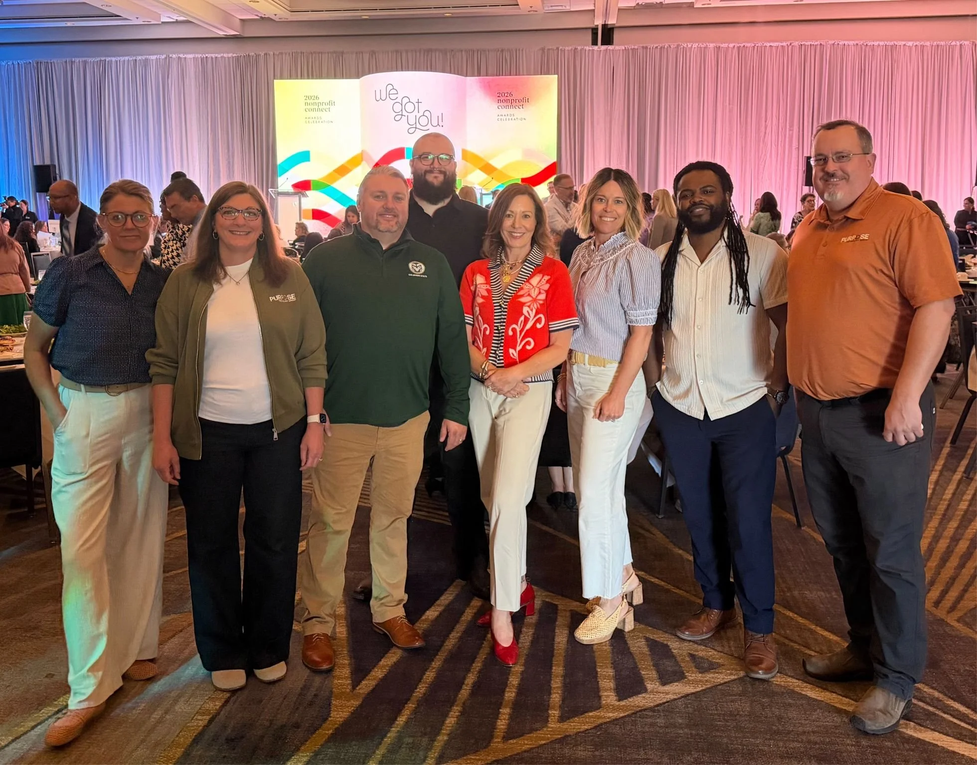 A diverse group of nine people standing together at an indoor event with a large colorful screen in the background that displays 'We got you!' and '2026 nonprofit connect awards celebration'.