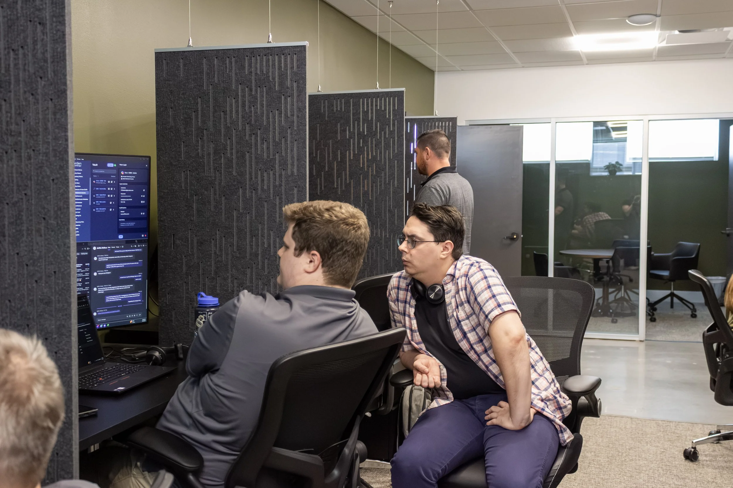 People working together in an office, focusing on a computer screen with code or a chat application. One person is seated, another is standing near a partition, and others are visible in the background through a glass wall.
