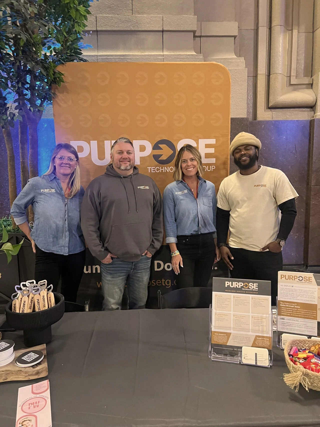 Four people standing behind a promotional table at an event, smiling at the camera. They are in front of a large banner that says 'PURPOSE' with additional text, and the table has branded items and informational displays.
