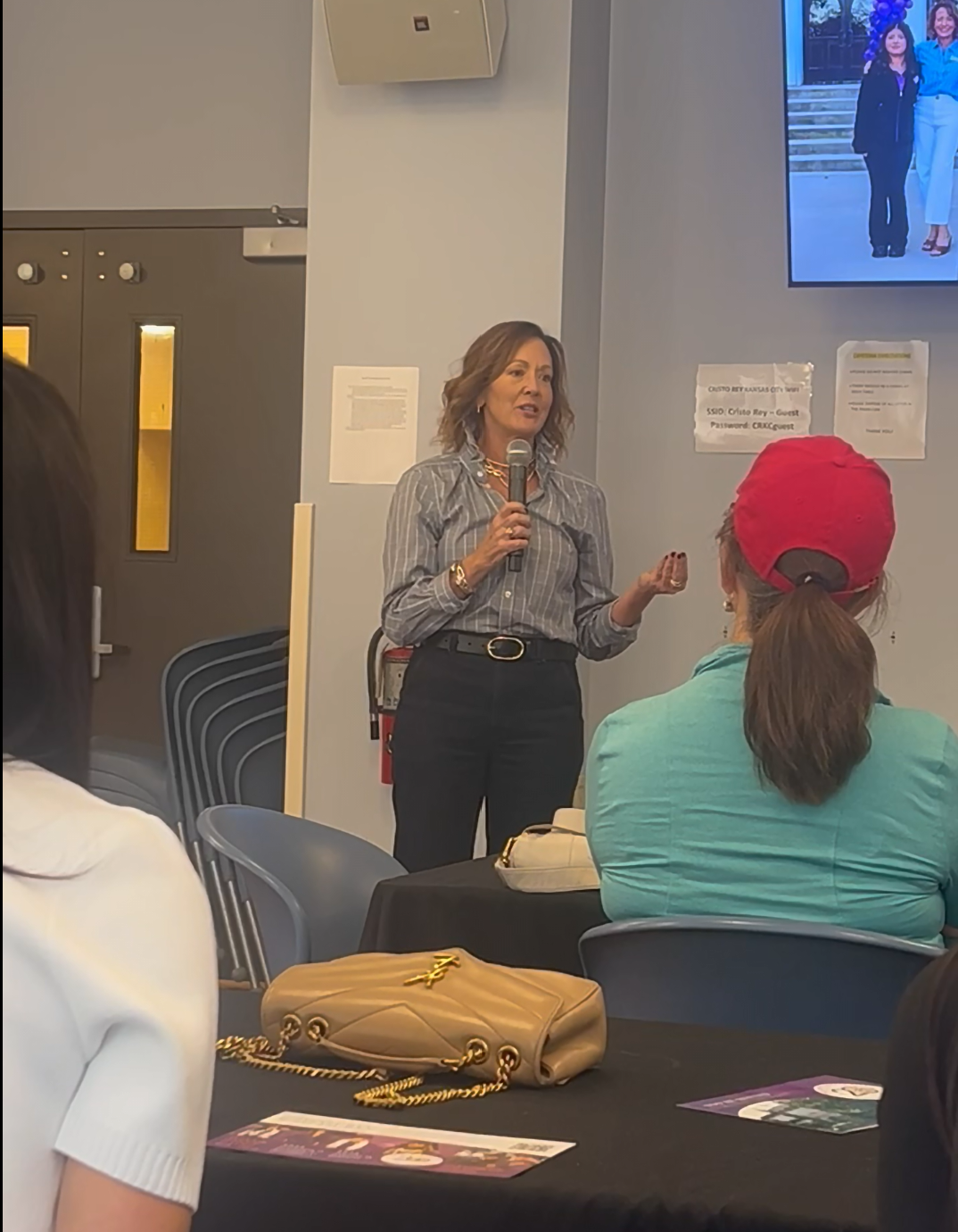 A woman is speaking into a microphone while addressing an audience in a room. She has shoulder-length brown hair and is wearing a gray striped shirt and black pants. There are people seated at tables, and a large screen behind her displaying an image