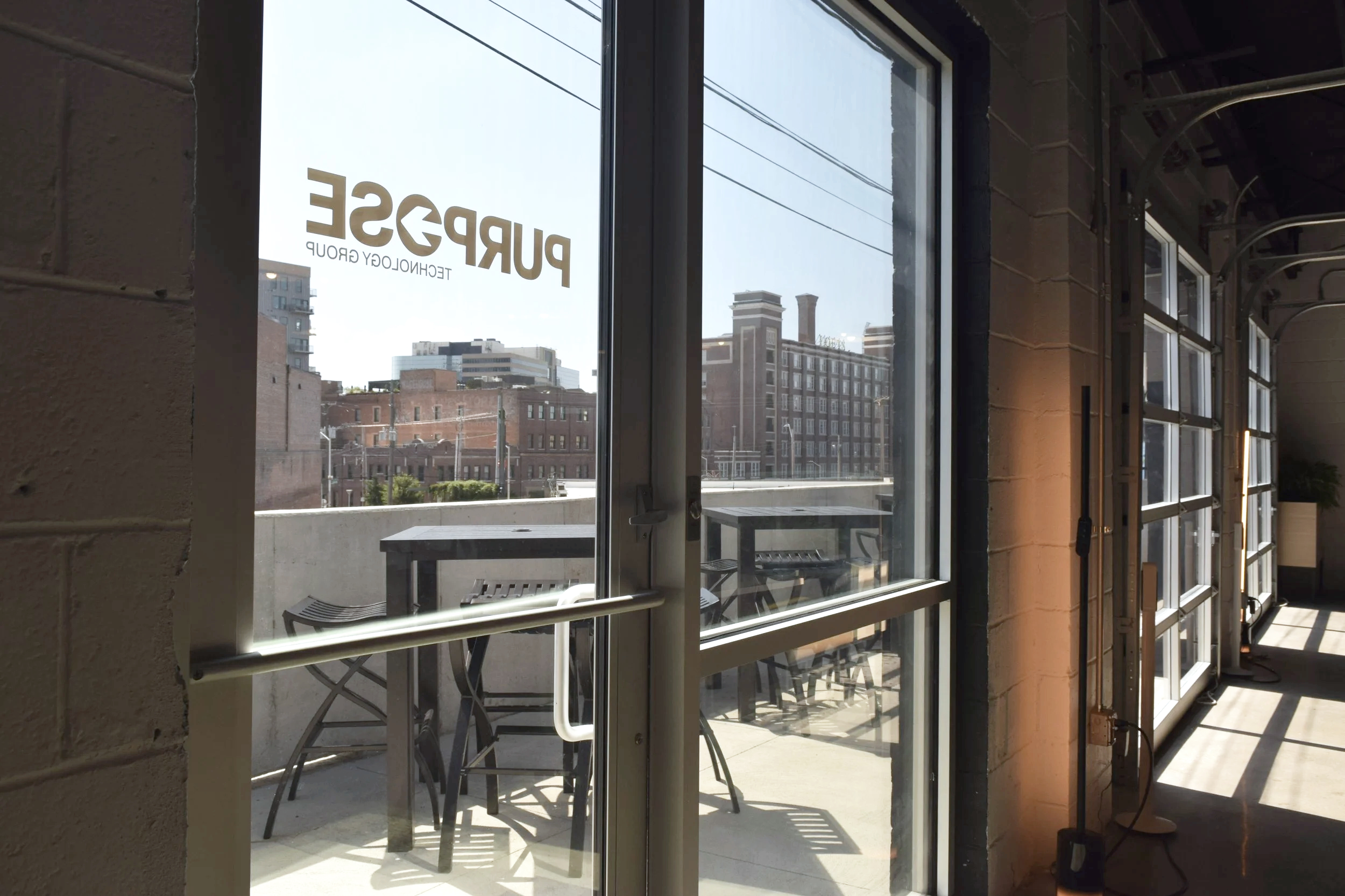 View through a glass door of an urban balcony with tables and chairs, city buildings, and power lines outside.
