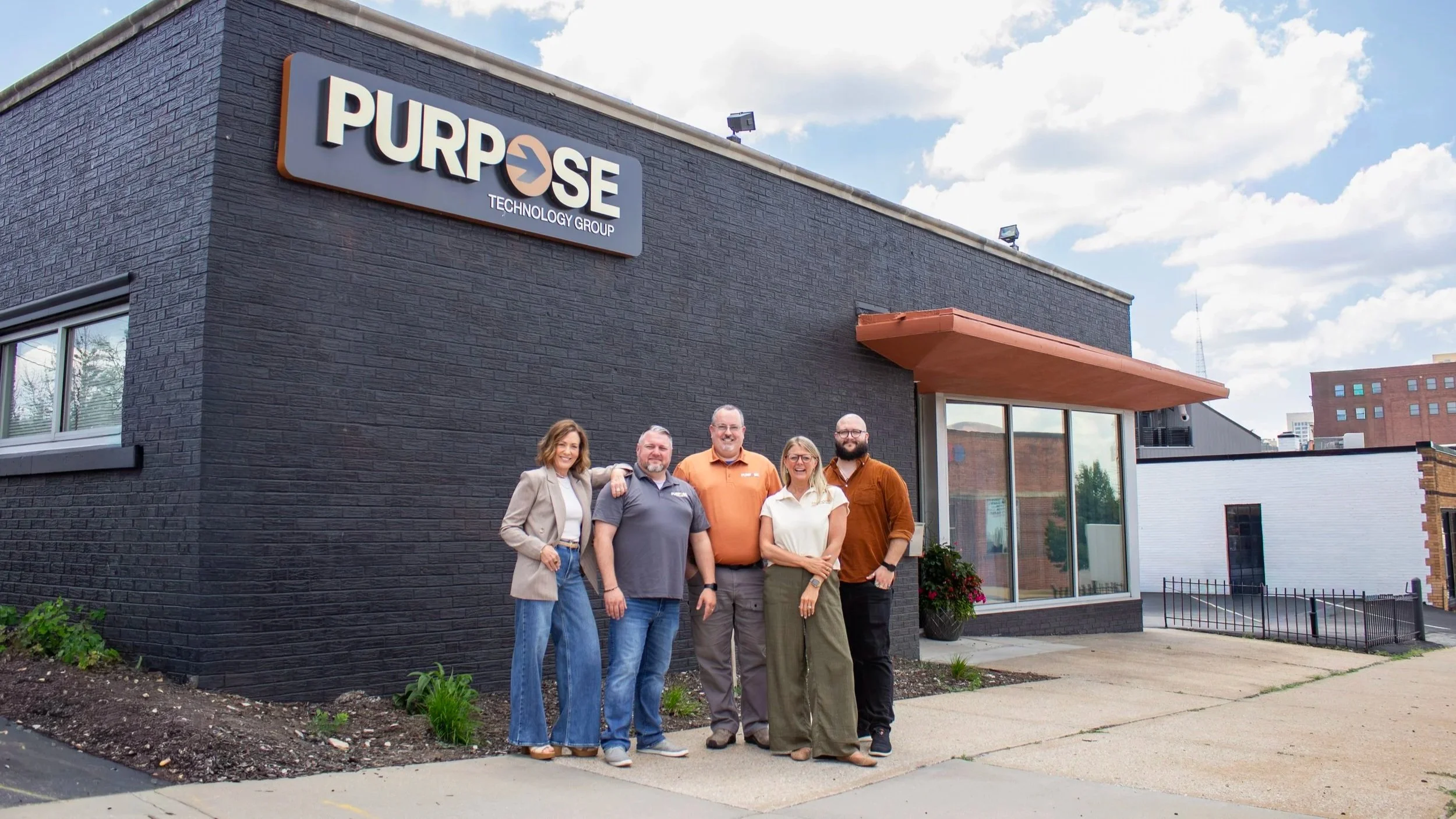A group of five people standing in front of a black brick building with a sign that reads 'PURPOSE TECHNOLOGY GROUP' on a partly cloudy day.