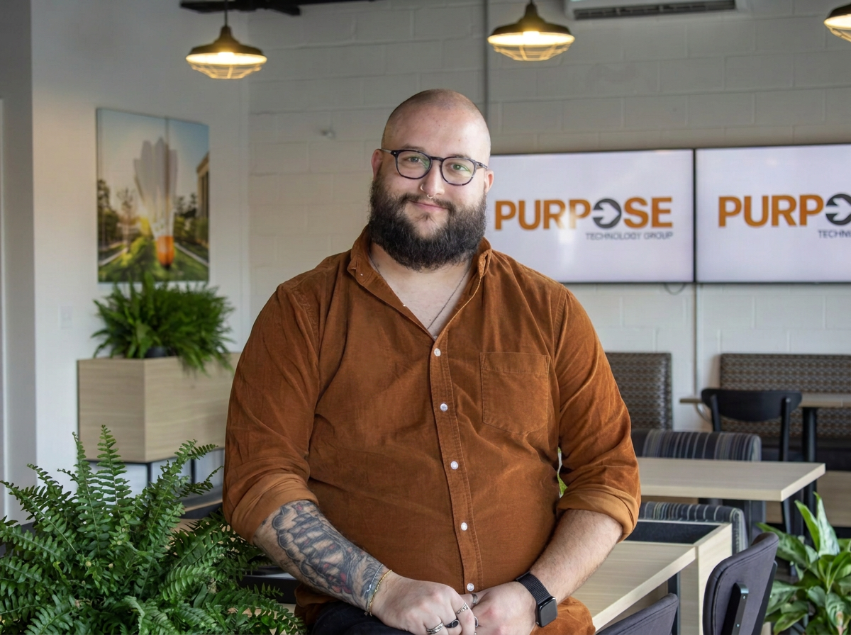 A bearded man with glasses, tattoos on his left arm, and wearing a brown shirt, stands in an office space with green plants, tables, chairs, and a wall with two screens displaying the word "PURPOSE".