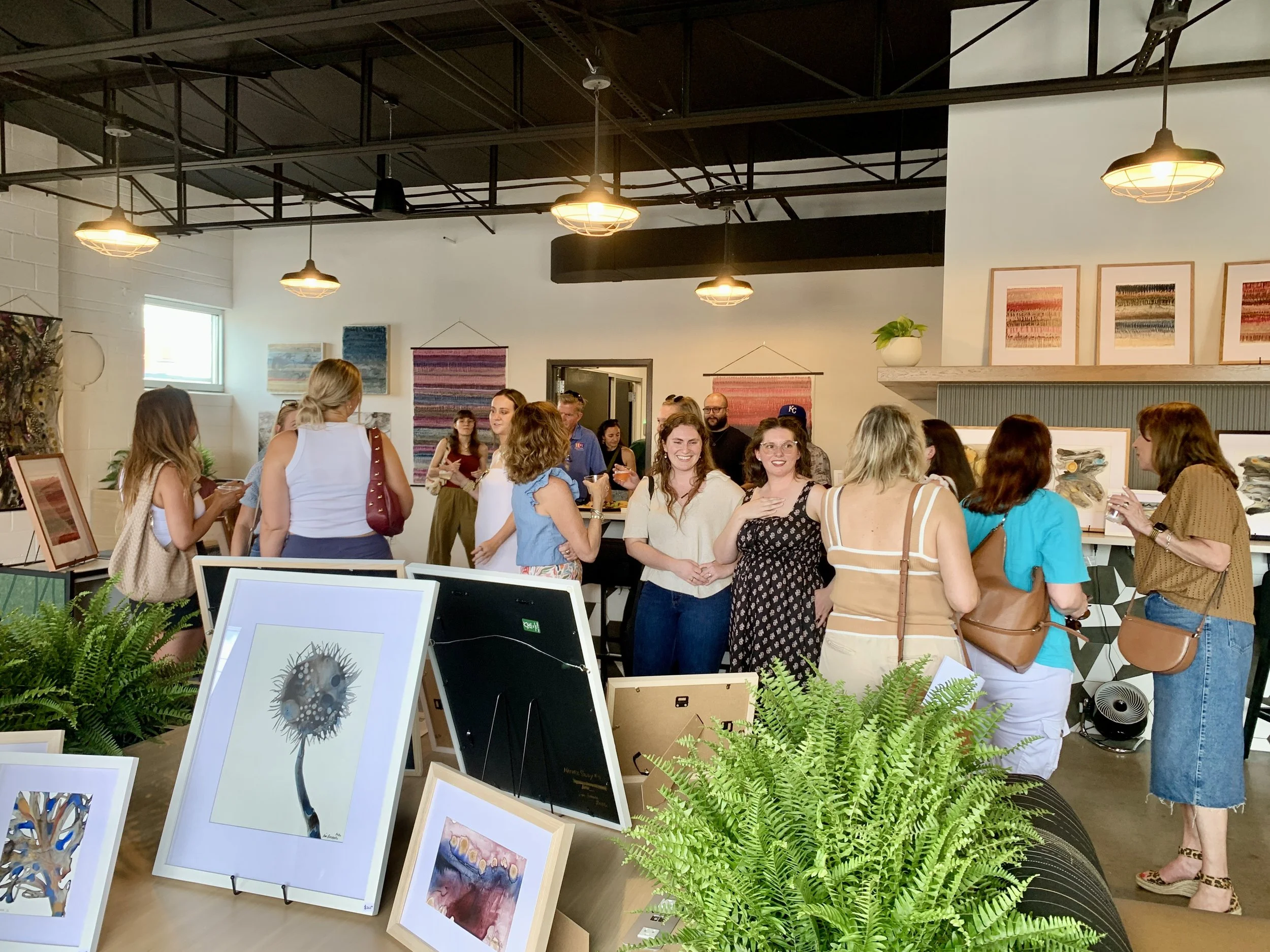 People gathering in an art gallery with paintings and artwork displayed on walls and on standalone frames, plants, and lighting fixtures overhead.