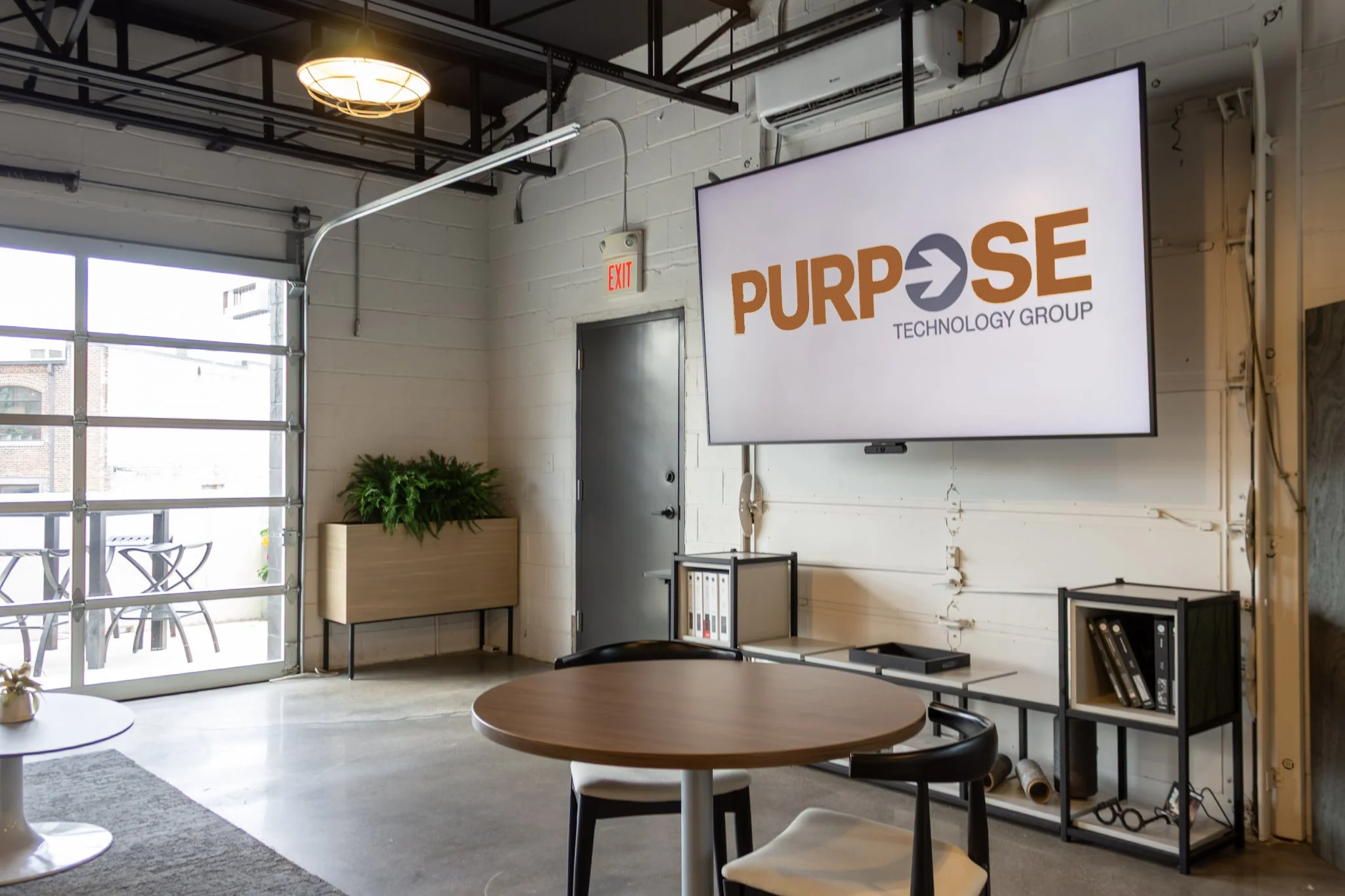 A photo of a modern office event space with garage doors and a screen that reads Purpose Technology Group.
