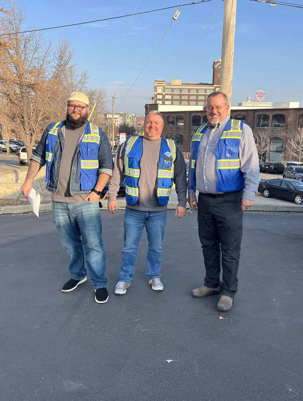 Three men standing outdoors on a paved area, wearing reflective safety vests, with a cityscape and parked cars in the background. All are smiling and dressed casually.