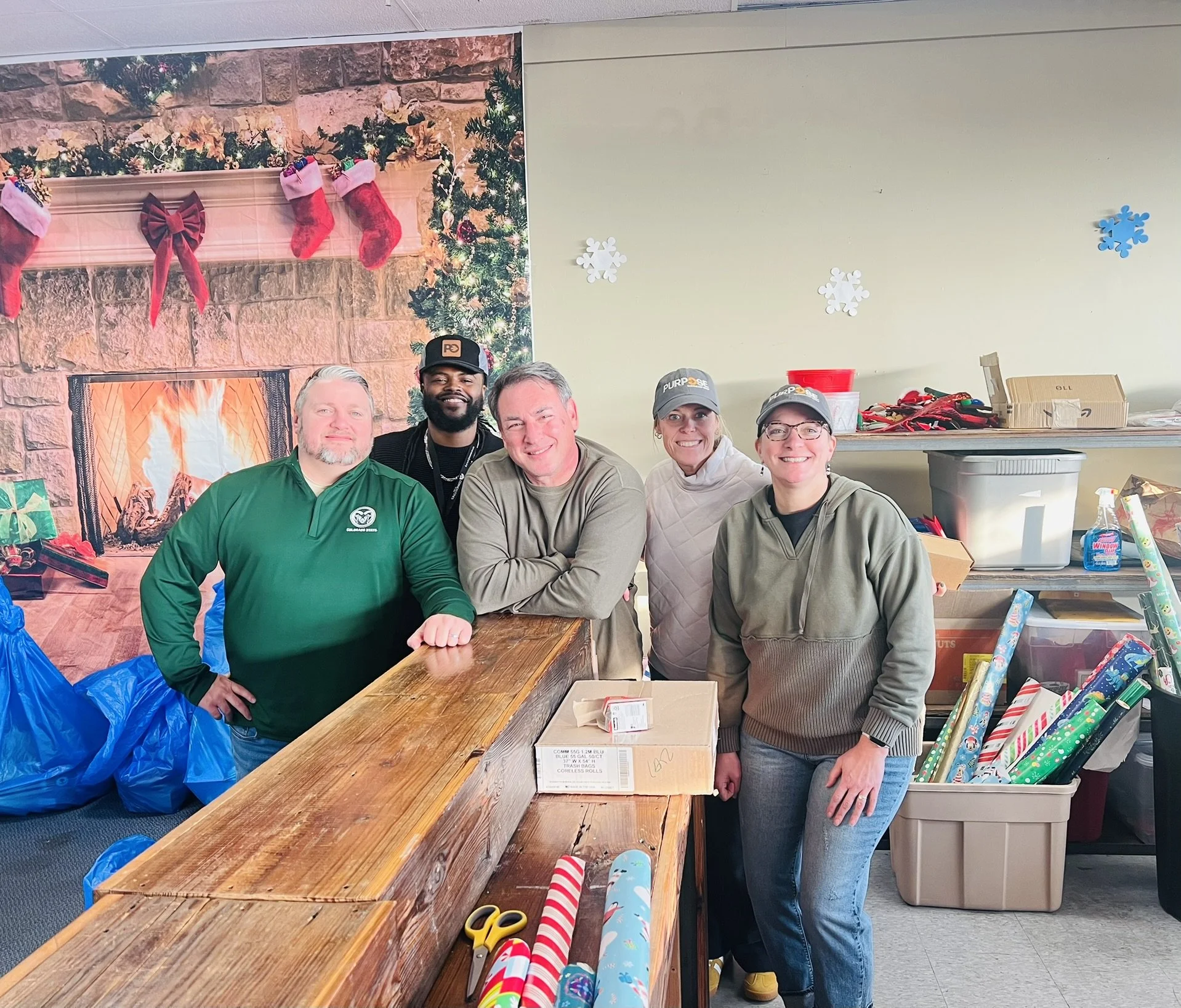 Group of five smiling people standing behind a wooden counter in a festive holiday setting with a fireplace, stockings, and Christmas decorations in the background. Wrapping paper and supplies are visible on and around the counter.
