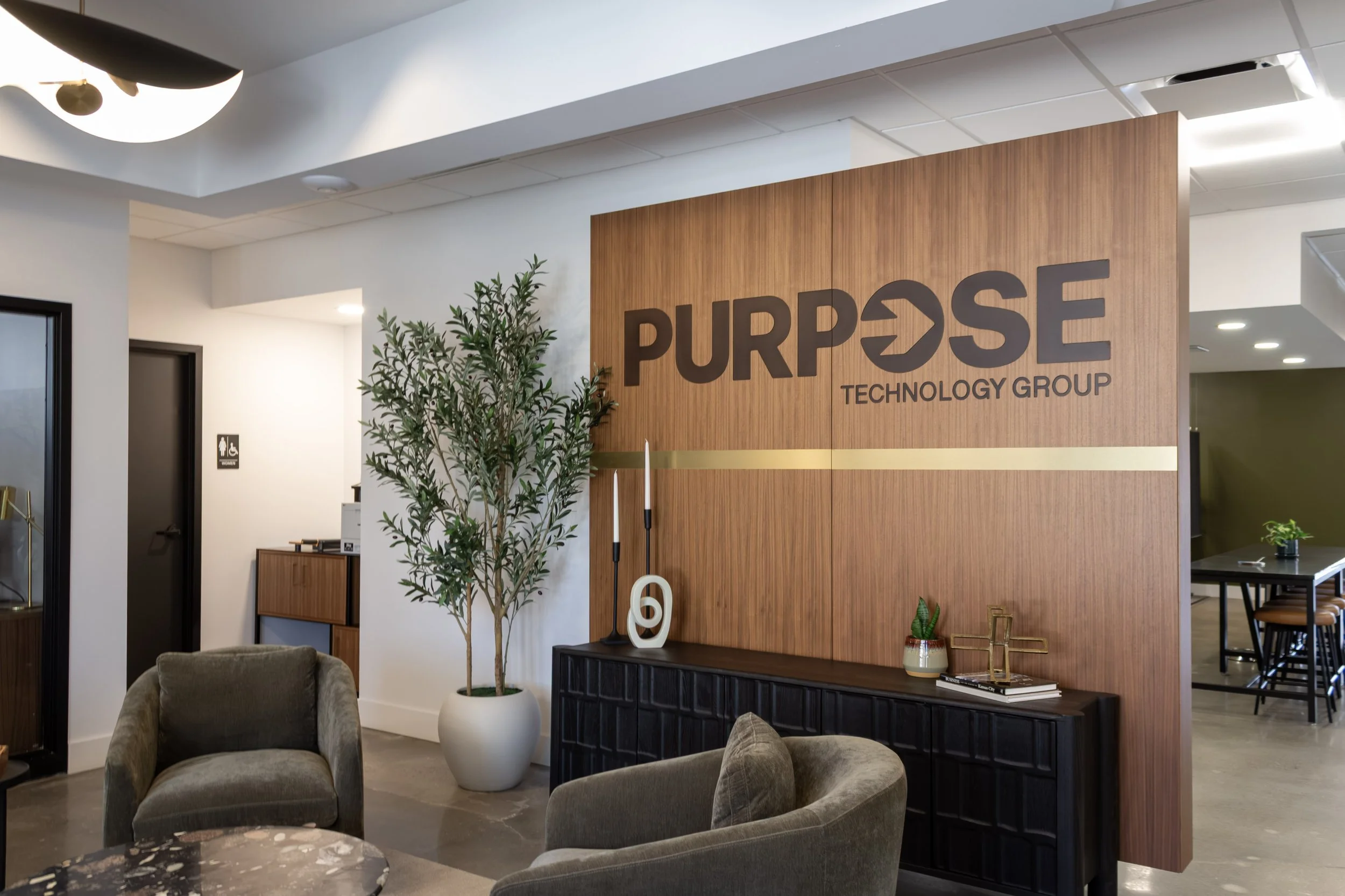 Interior of an office reception area featuring a large wooden wall with the company logo 'PURPOSE TECHNOLOGY GROUP', decorative plants, a black sideboard with decor items, and comfortable seating.
