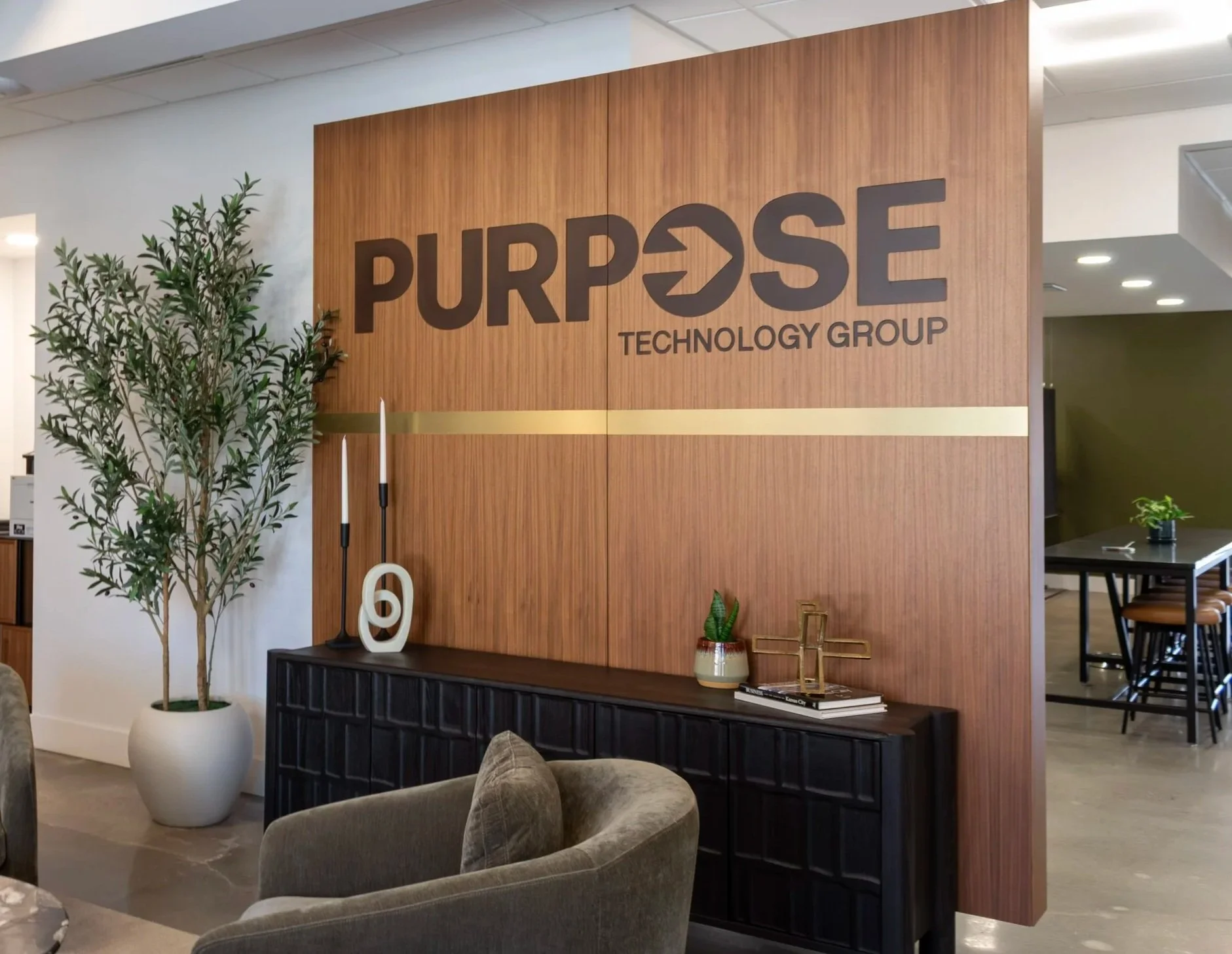 Interior of an office reception area featuring a large wooden wall with the company logo 'PURPOSE TECHNOLOGY GROUP', decorative plants, a black sideboard with decor items, and comfortable seating.