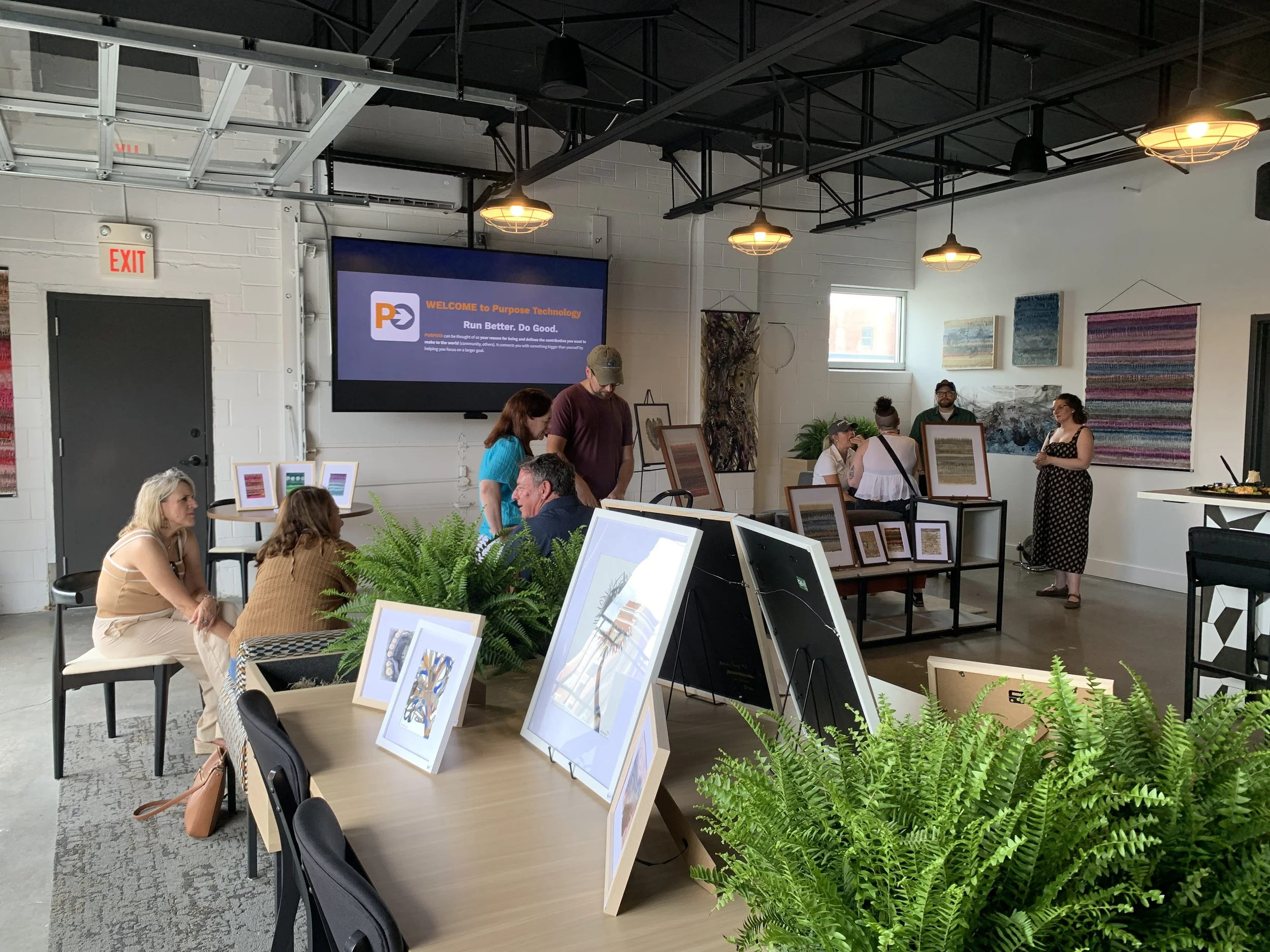 An art gallery or exhibition space with artworks displayed on tables and walls, several people viewing and discussing art, and a large screen with a welcome message at the back.