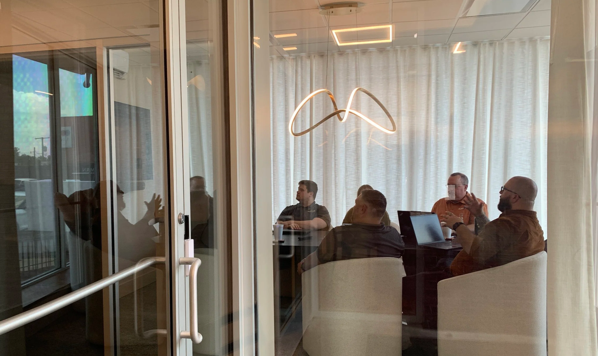 People sitting at a conference table in a room with large windows and curtains, engaging in a discussion. Modern lighting fixture hangs from the ceiling.