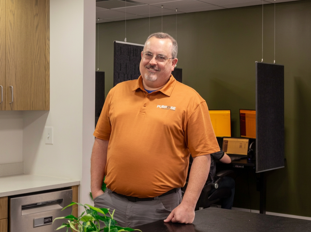 Man smiling in an office or technology workspace, wearing an orange polo shirt with a 'PURPOSE' logo, standing with one hand on his hip.