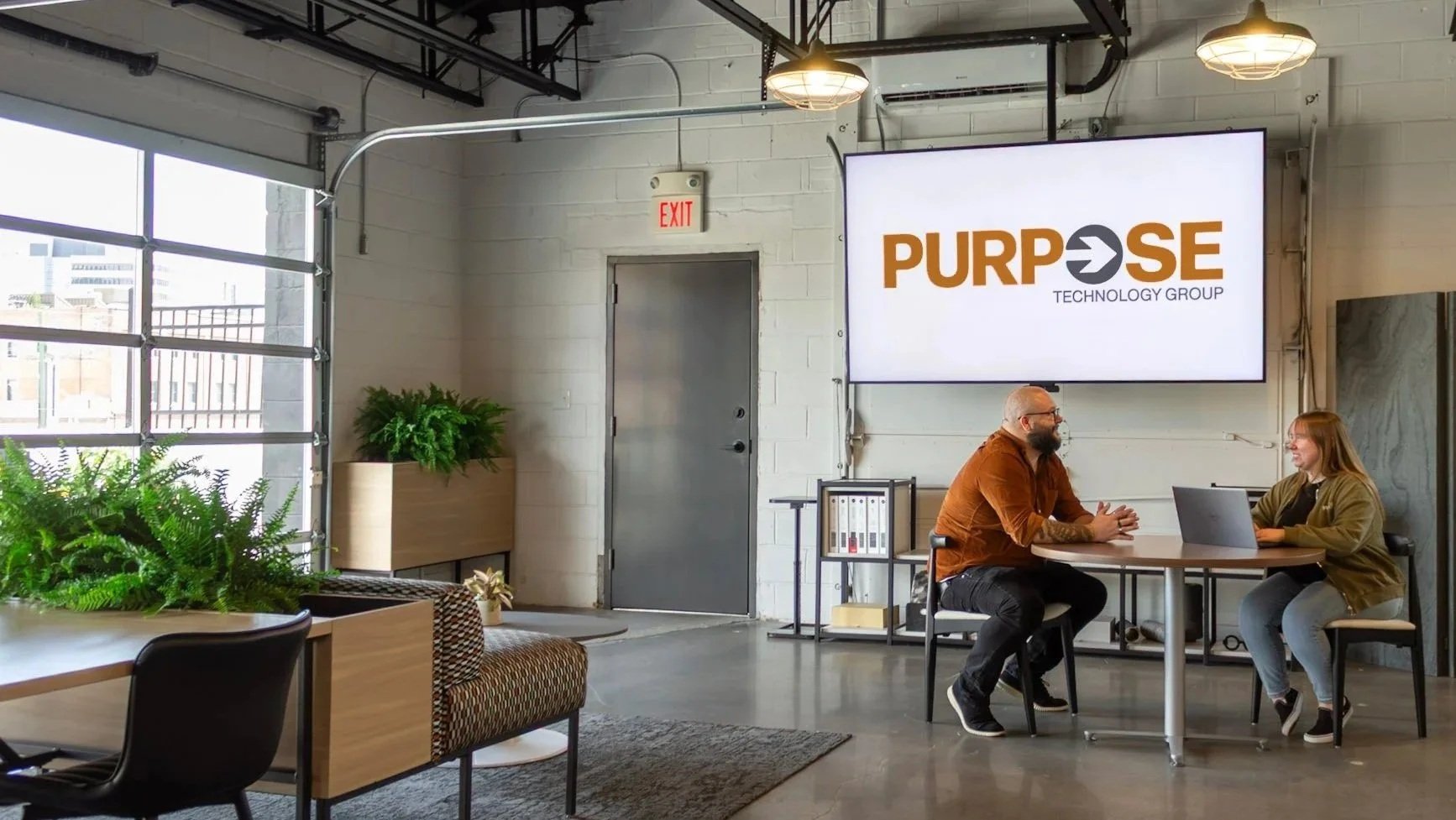 Two people sitting at a round table in a modern office space, engaged in conversation, with a large screen behind them displaying the logo for PURPOSE Technology Group.