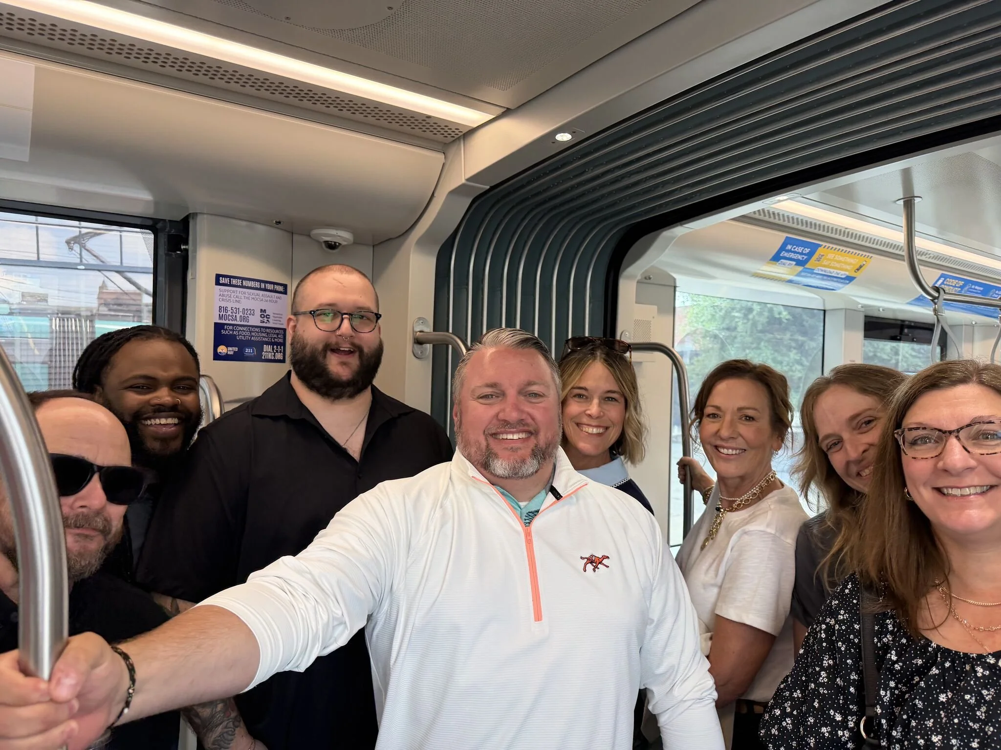 A group of smiling people inside a train or subway, taking a selfie together. The group includes men and women of diverse ethnicities and ages, with some wearing glasses, and one person has a beard. The background shows parts of the train interior, i