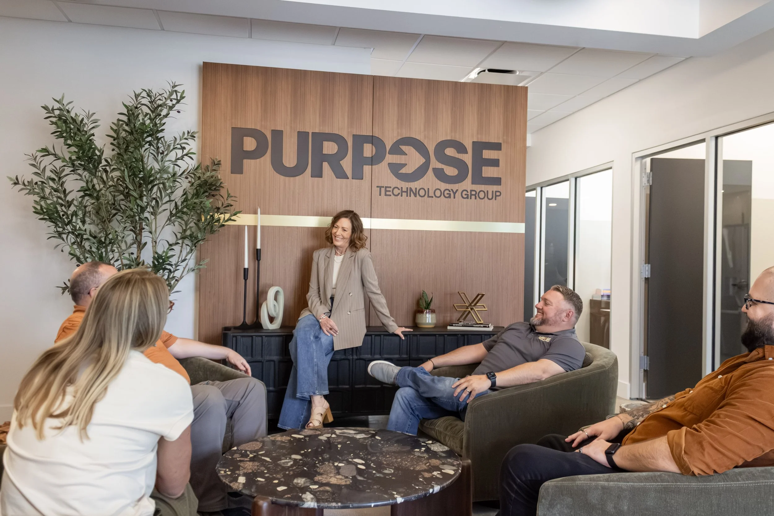 Group of people sitting and standing in a modern office space, engaging in conversation, with a woman in a tan blazer smiling and leaning against a black sideboard, and a large sign reading 'PURPOSE TECHNOLOGY GROUP' on the wooden wall behind her.
