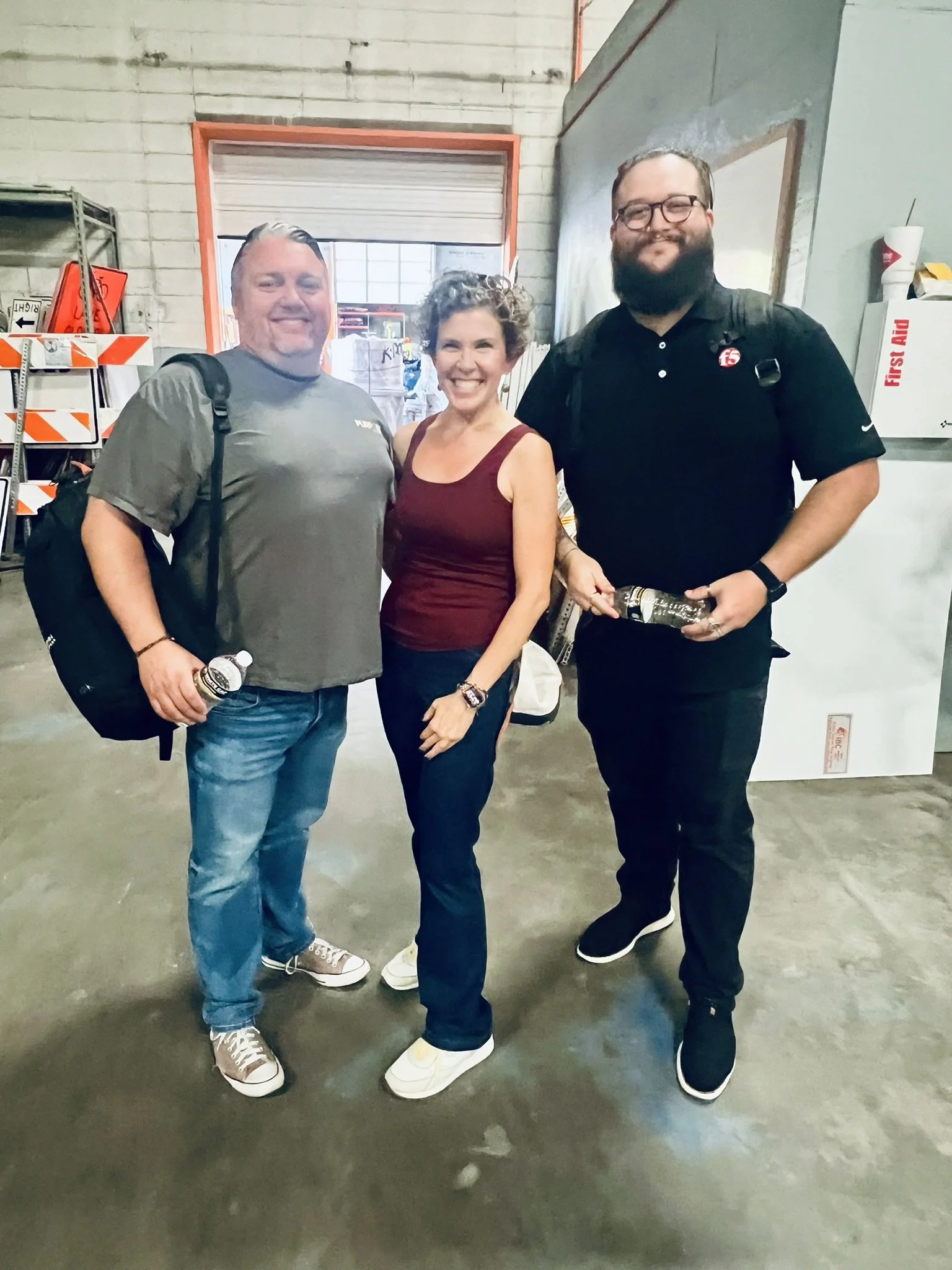 Three people standing together indoors, smiling at the camera, with a warehouse-like background including shelves, a window, and a whiteboard with some kitchen items and a fire extinguisher.