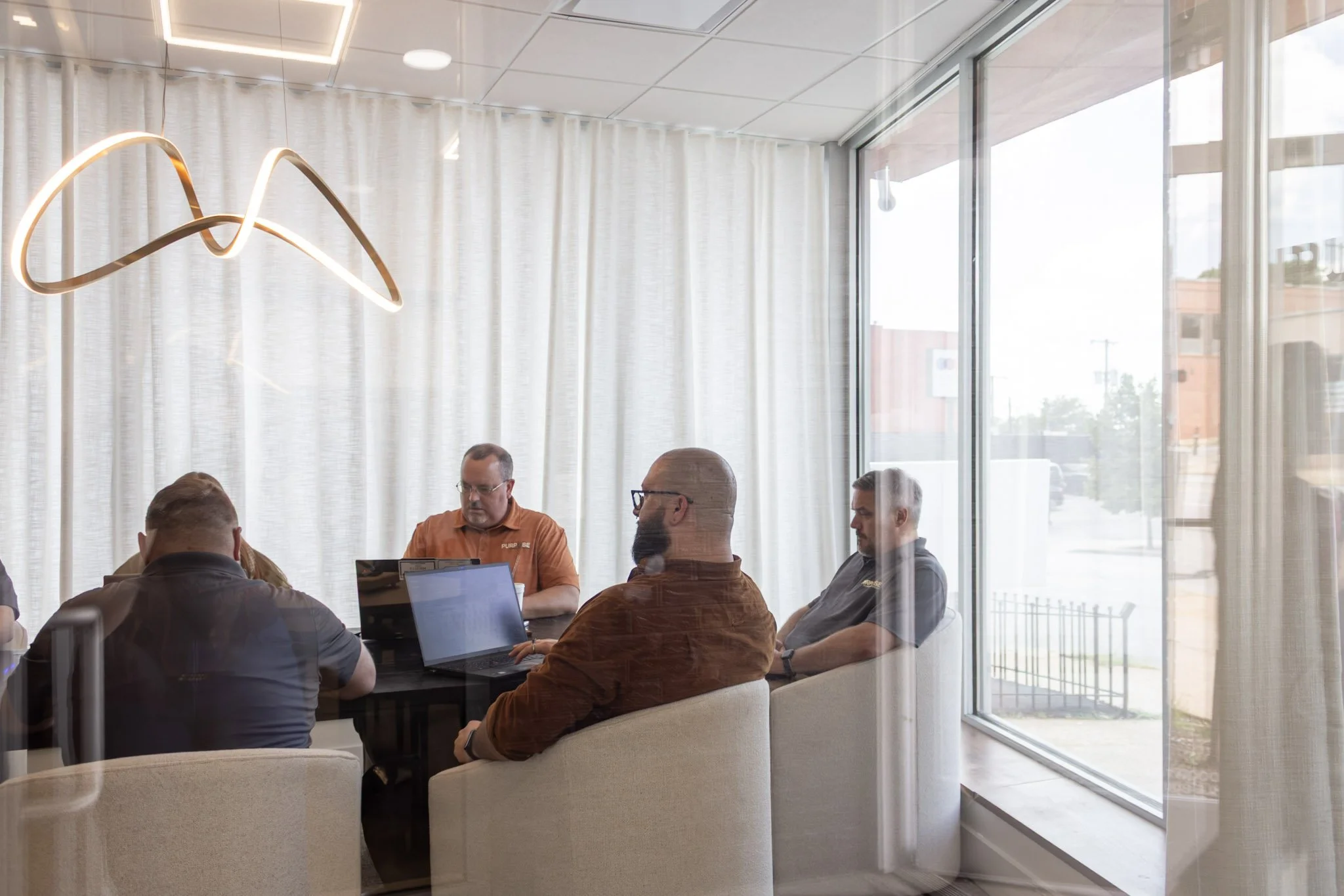 A group of men sitting around a table with laptops and documents inside a modern conference room with large windows and white curtains.