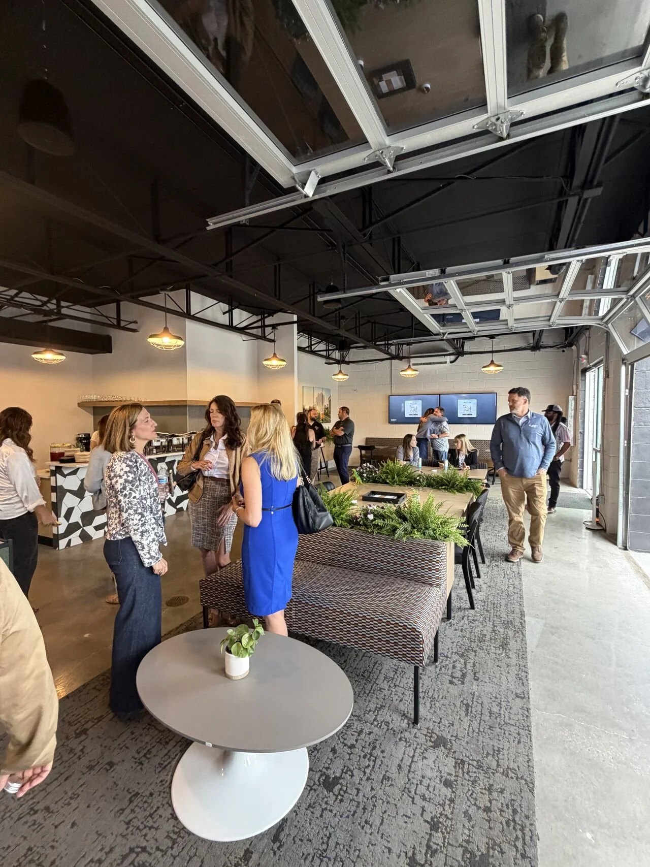 Indoor gathering with people talking and networking in a modern, open-concept space with a high ceiling, black and white decor, and digital screens on the wall.