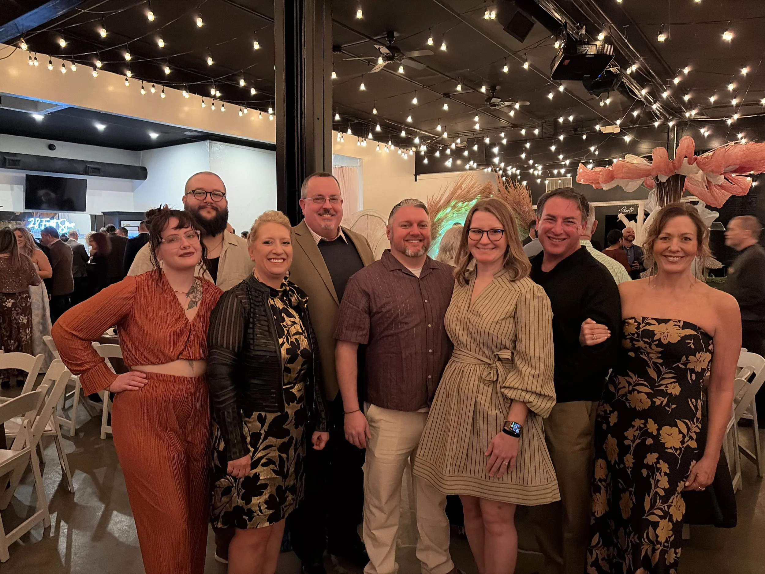 A group of nine people smiling and posing together at an indoor event with string lights overhead.