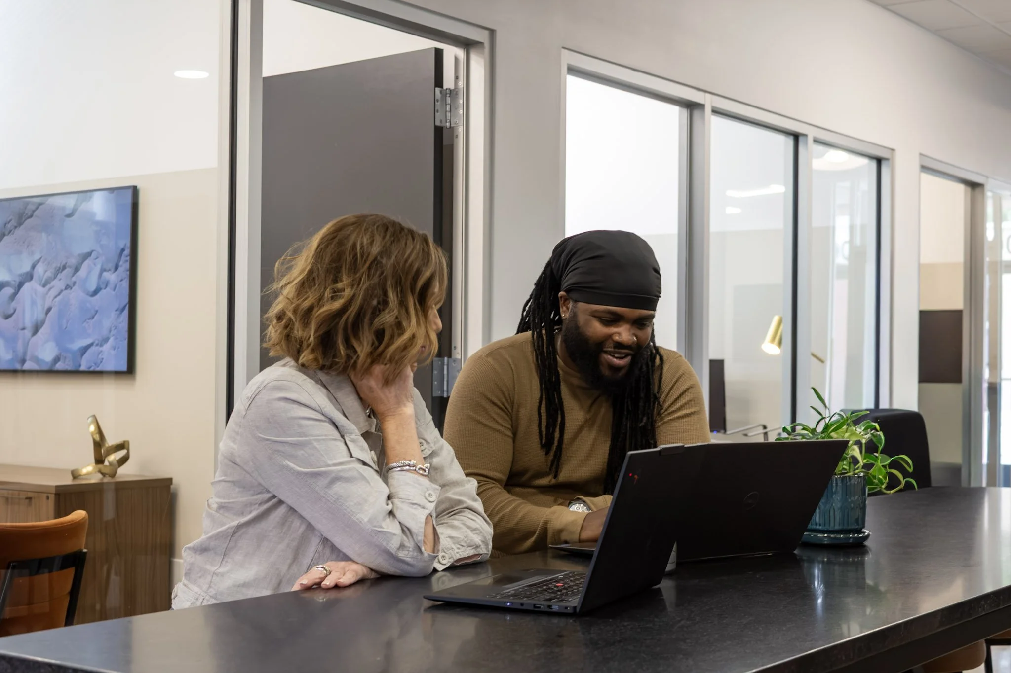 Two people sit at a counter and look at a laptop, engaged in a discussion in a modern office space with glass walls and a television on the wall.