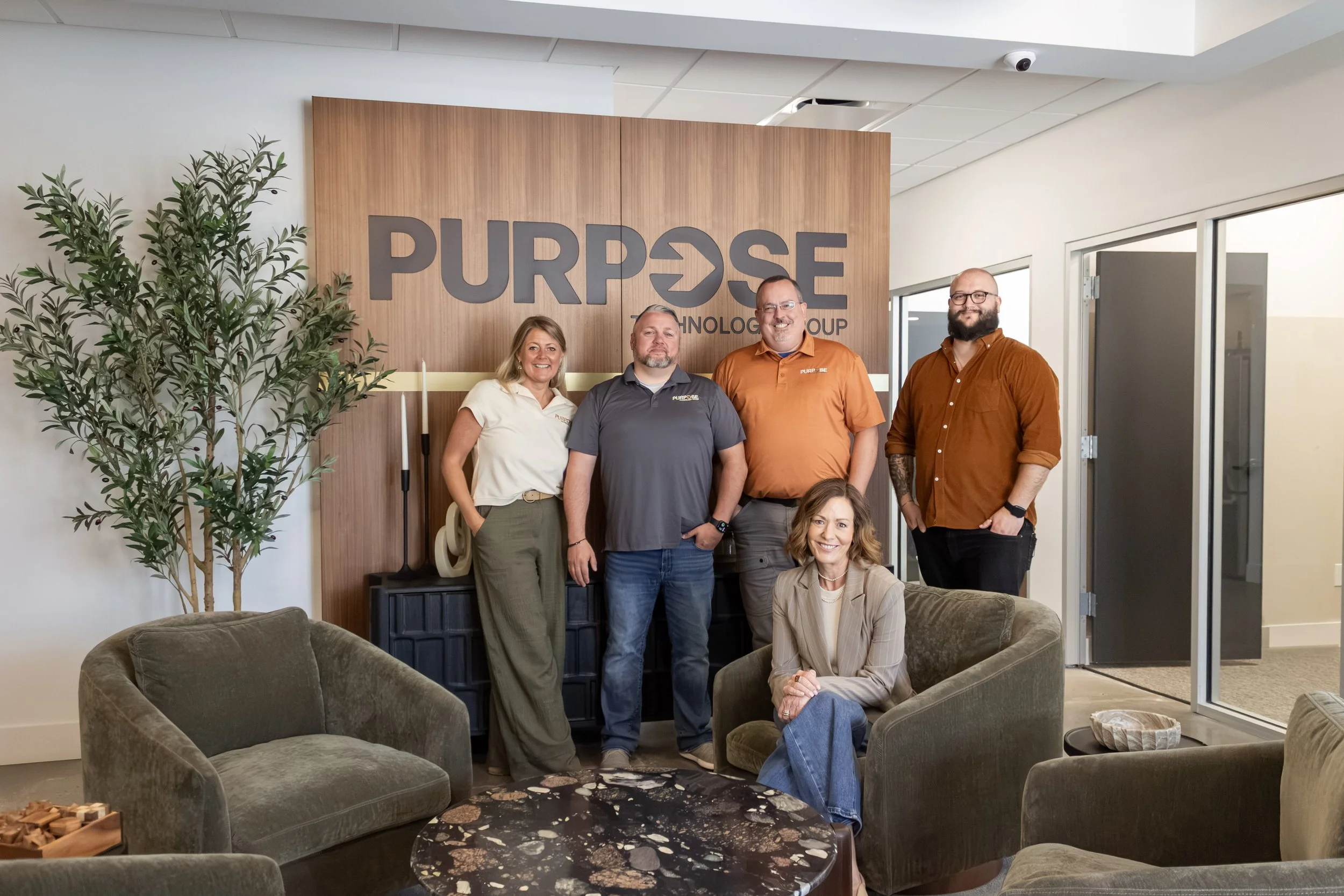 A group of five people standing and one woman sitting in an office lobby, in front of a wooden wall with the company logo 'PURPOSE Technology Group.' They are smiling and dressed in business casual attire. The lobby has a sofa, armchairs, a coffee table, and a large green plant.