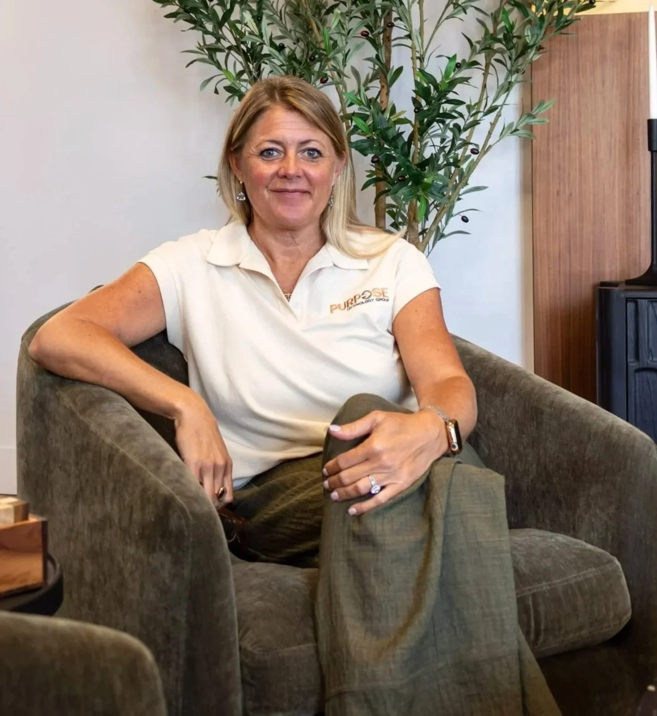 A woman with blonde hair sitting on a brown velvet armchair, wearing a white polo shirt with a logo, and olive-green pants, smiling at the camera with a large green plant behind her.