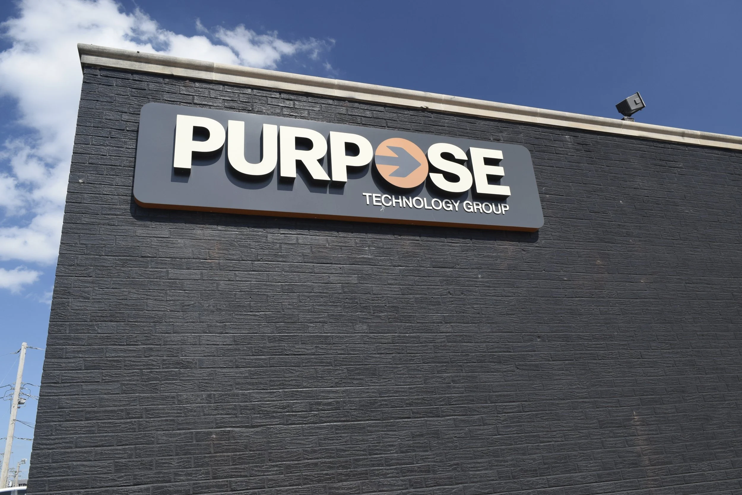 Front of the Purpose Technology Group Building in Kansas City, the building sign is the logo that reads 'Purpose Technology Group' mounted on a dark brick wall, under a partly cloudy sky.