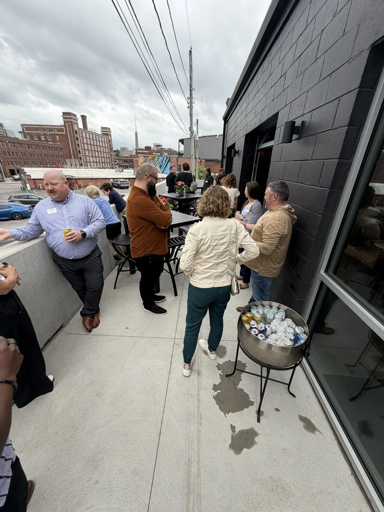 People gathered on a rooftop terrace with a black brick wall, drinking beverages, and socializing under a cloudy sky. A table with cans of drinks and foil-wrapped items is visible. The city skyline with buildings and power lines can be seen in the ba