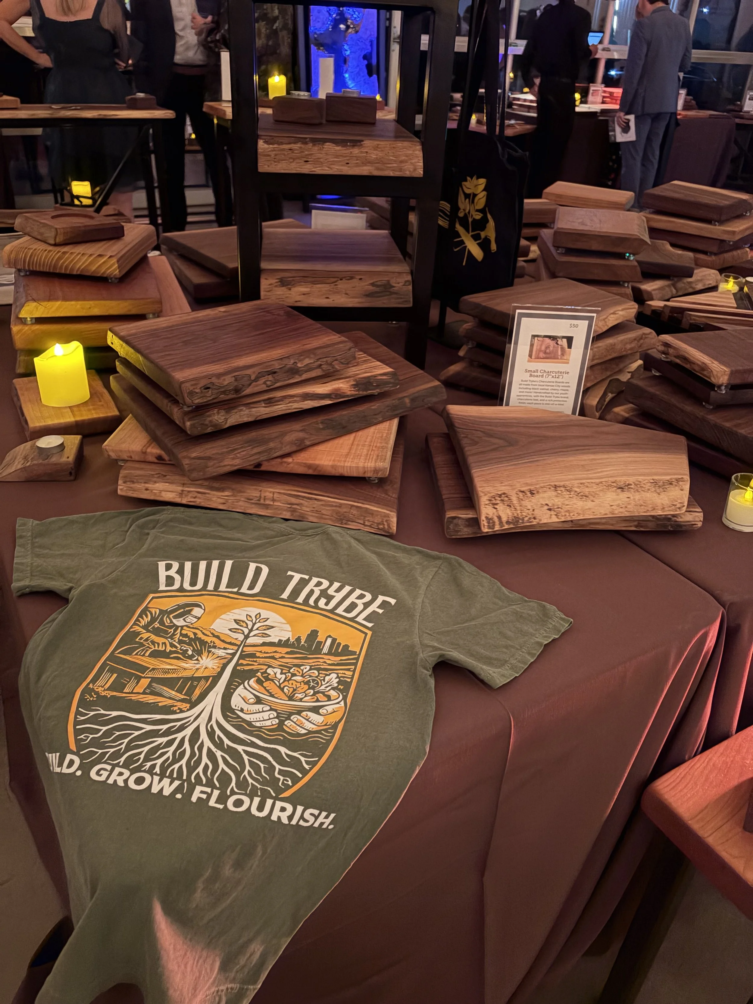 A display of wooden chopping boards on a table at an event, with some people in the background and a small charcuterie board sign.