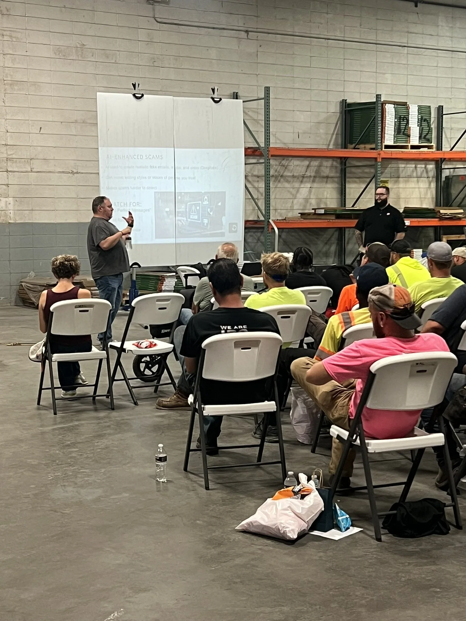 A man is giving a presentation to a seated audience inside a warehouse or large industrial space. The screen displays a slide titled 'AI-Enhanced Scams'. The audience includes people wearing safety vests and casual clothing, and there is a table with