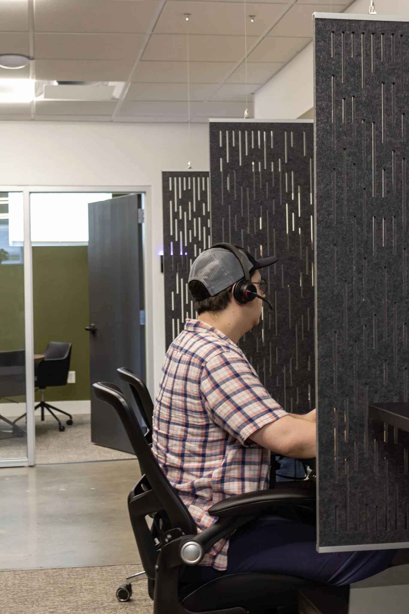 A man wearing a plaid shirt, black glasses, and headphones, working at a computer in an office with partition walls.