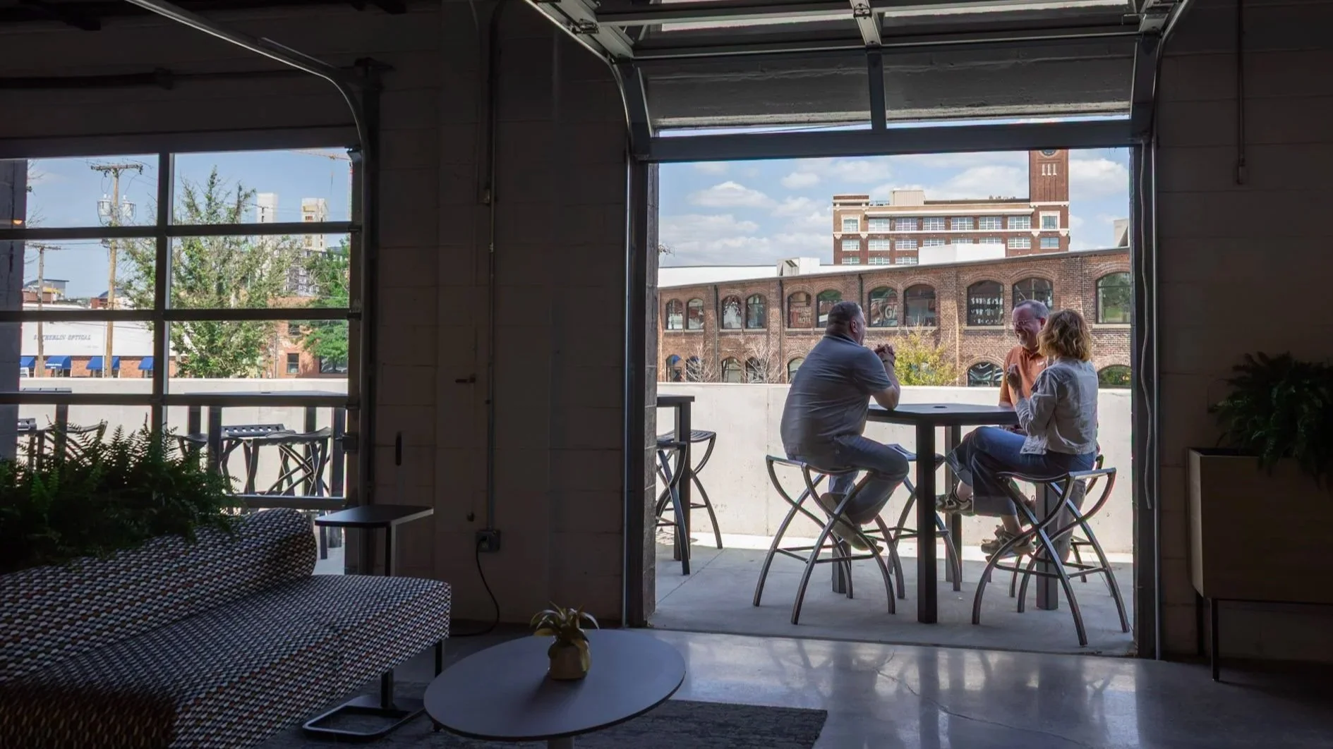 Three people sitting around a small table on an outdoor balcony, talking, with urban buildings in the background.