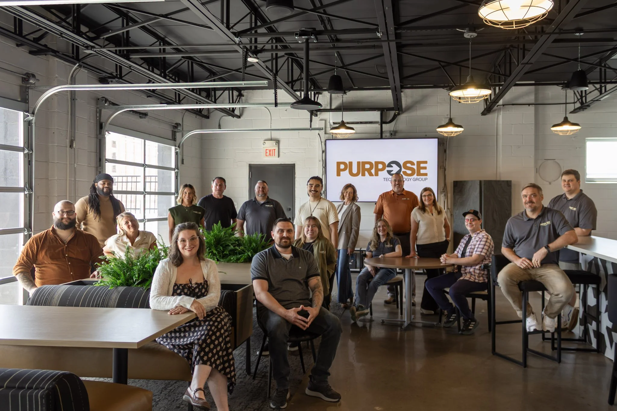A diverse group of people gathered in a modern, industrial-style office space with large windows, hanging lights, and a large screen displaying the word 'PURPOSE' along with 'TECHNOLOGY GROUP' underneath. The group is smiling and posing for the photo.