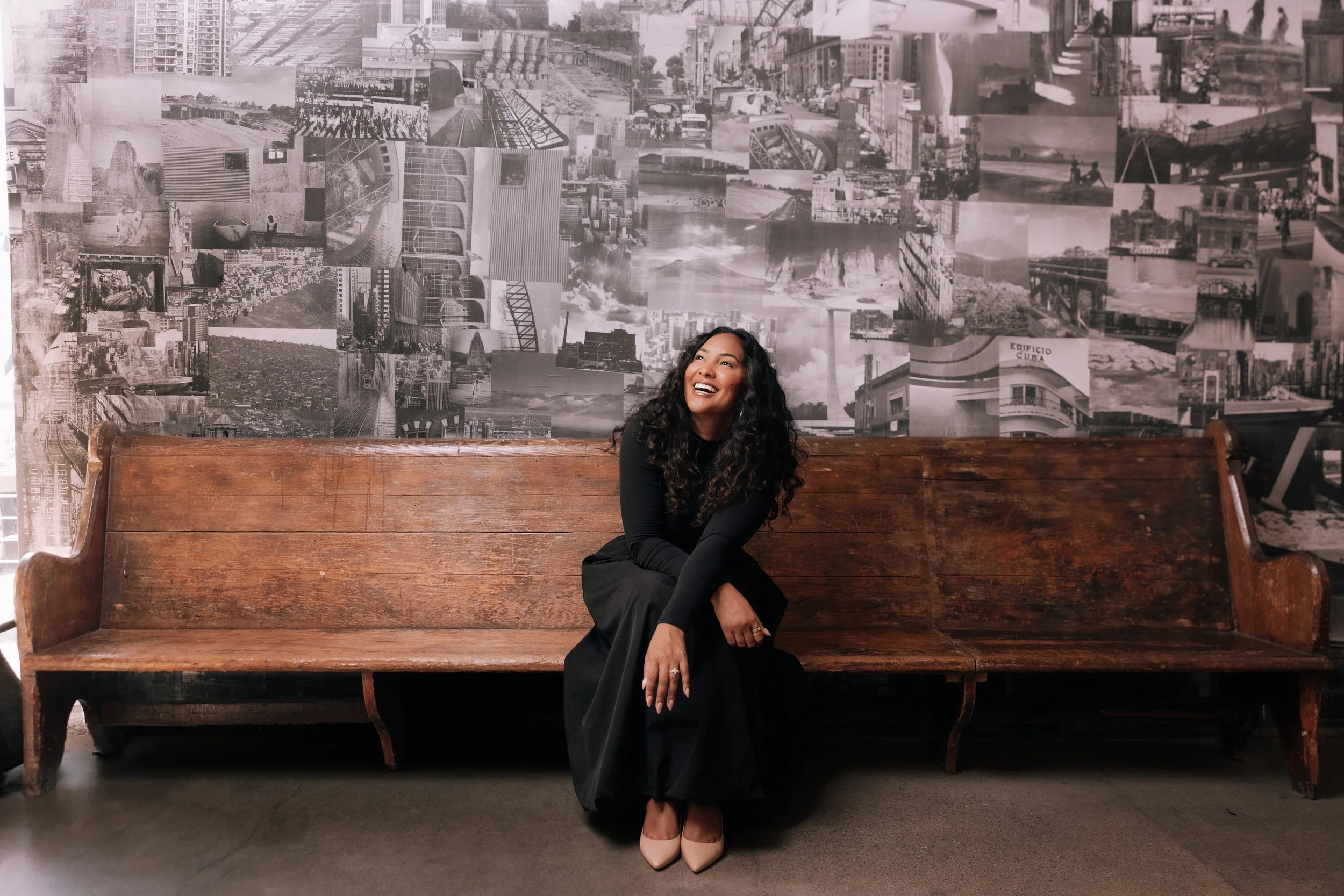 A woman with curly black hair sitting on a wooden bench, smiling, in front of a collage of black and white city photographs on the wall, wearing a black dress and tan high heels.