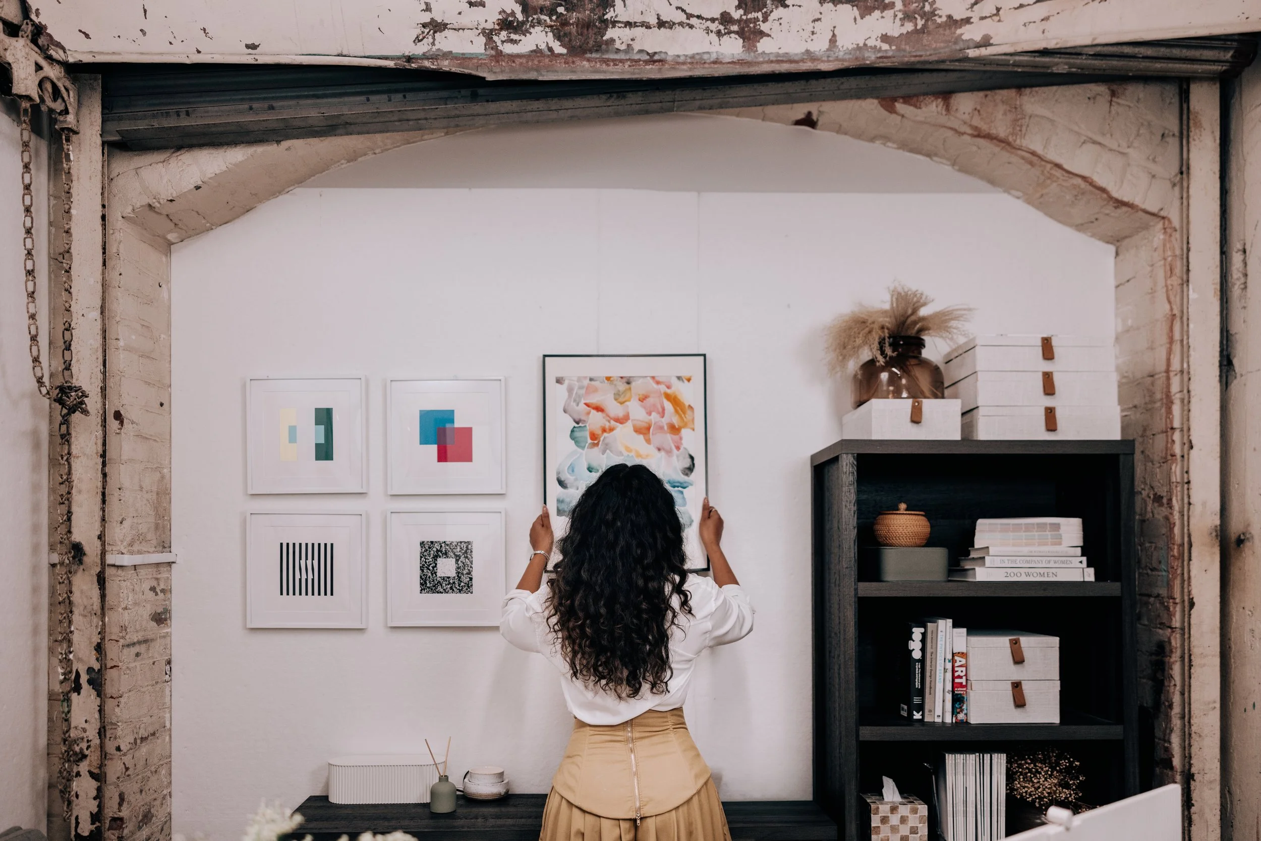A woman with long curly hair hanging a photo of framed watercolor artwork on a white wall in a stylish, industrial-style studio with exposed brick and a black bookshelf.