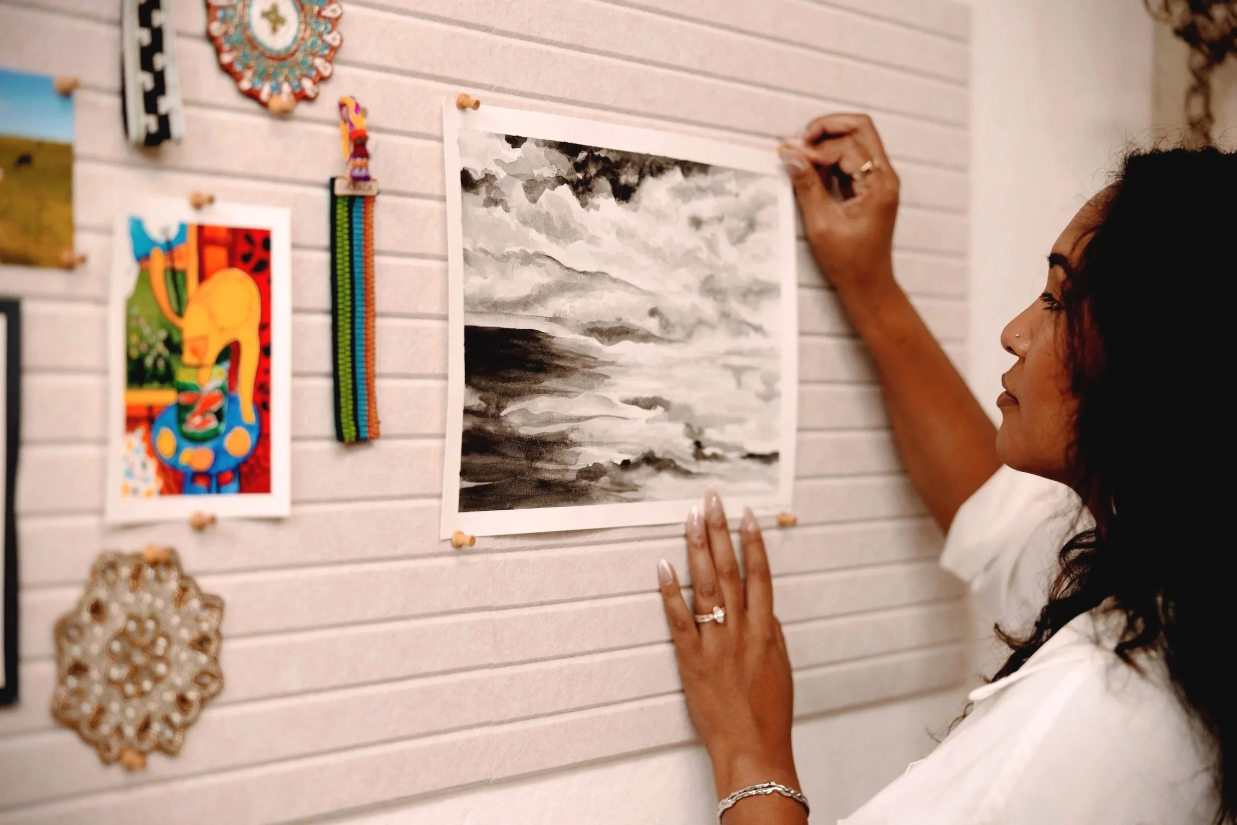 A woman hanging a black and white watercolor painting of a cloudy sky and sea on a wall. The wall has pinboard panels with various small decorations, including colorful and beaded artwork.