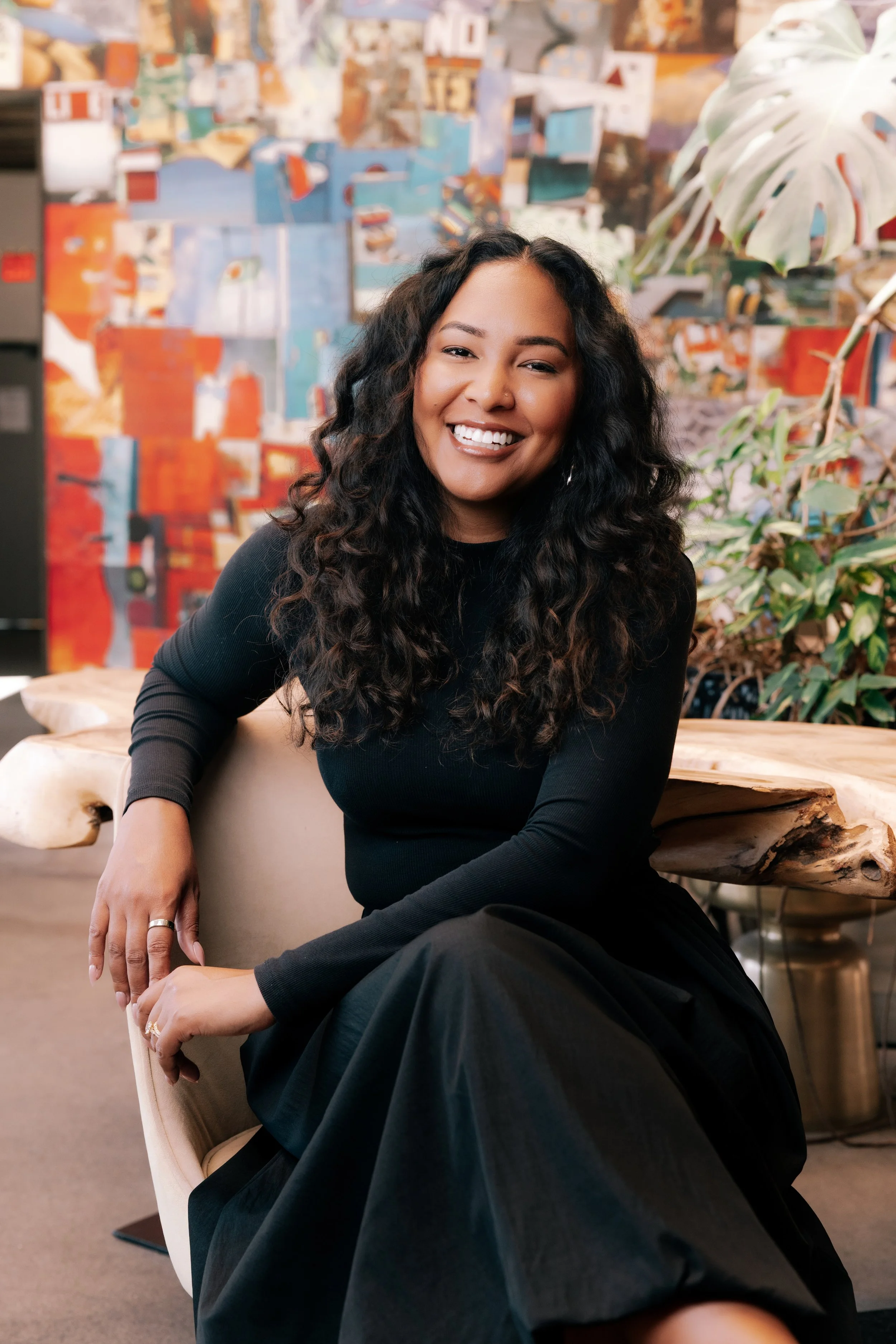A woman with long curly black hair and a smile, wearing a black long-sleeve top, sitting in a modern indoor setting with colorful abstract artwork on the wall and green plants in the background.