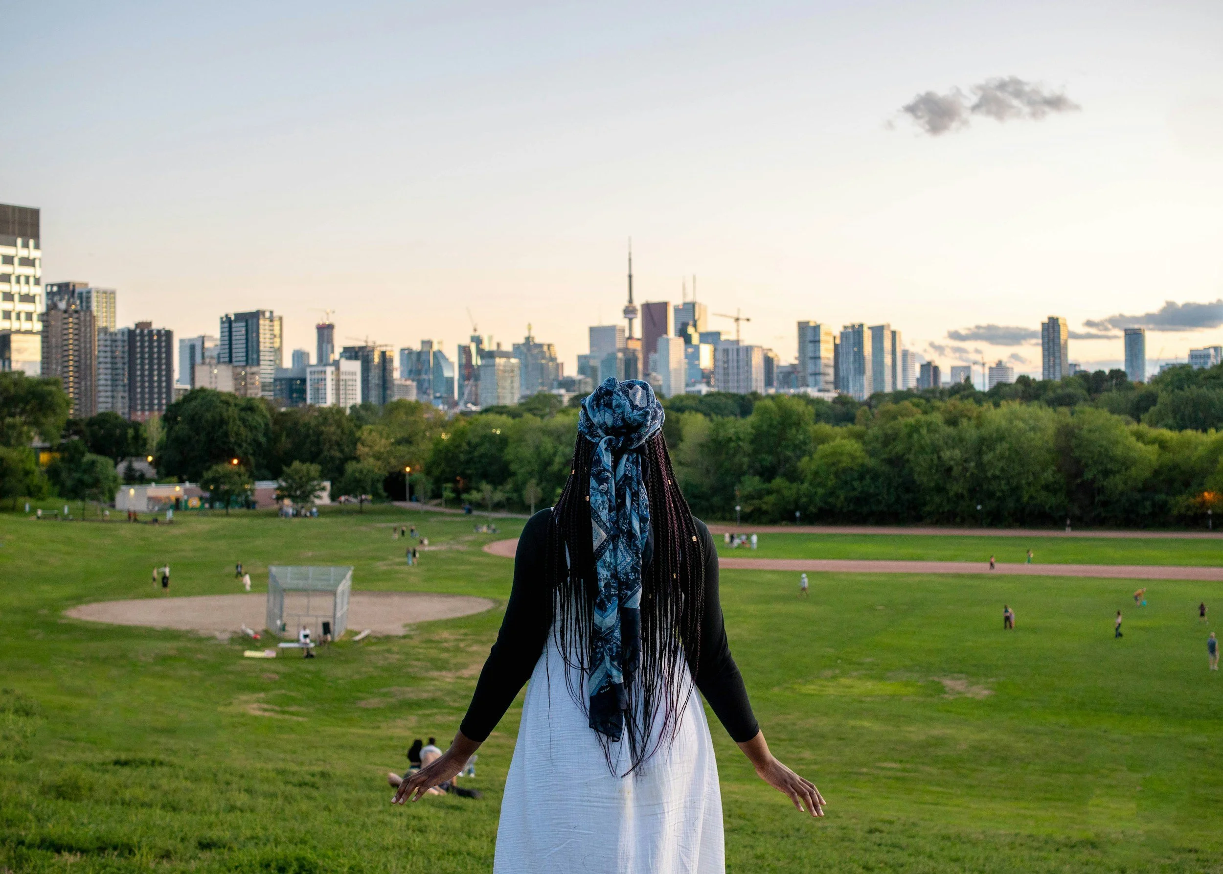 A woman with long braided hair, wearing a black top, white skirt, and a blue patterned scarf, practicing yoga or meditation in a park with a city skyline in the background during sunset.