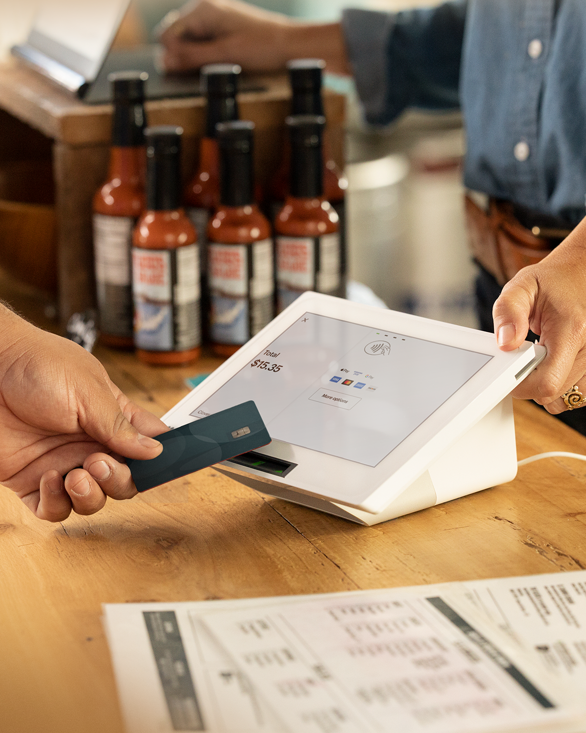 Person making a contactless payment with a credit card on a point-of-sale touchscreen device, with bottles of hot sauce in the background.