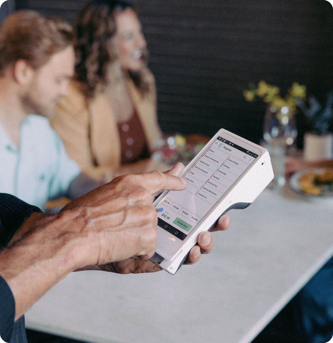 A person uses a handheld point-of-sale device at a restaurant table with three seated women in the background, with plates of food and a vase of flowers on the table.