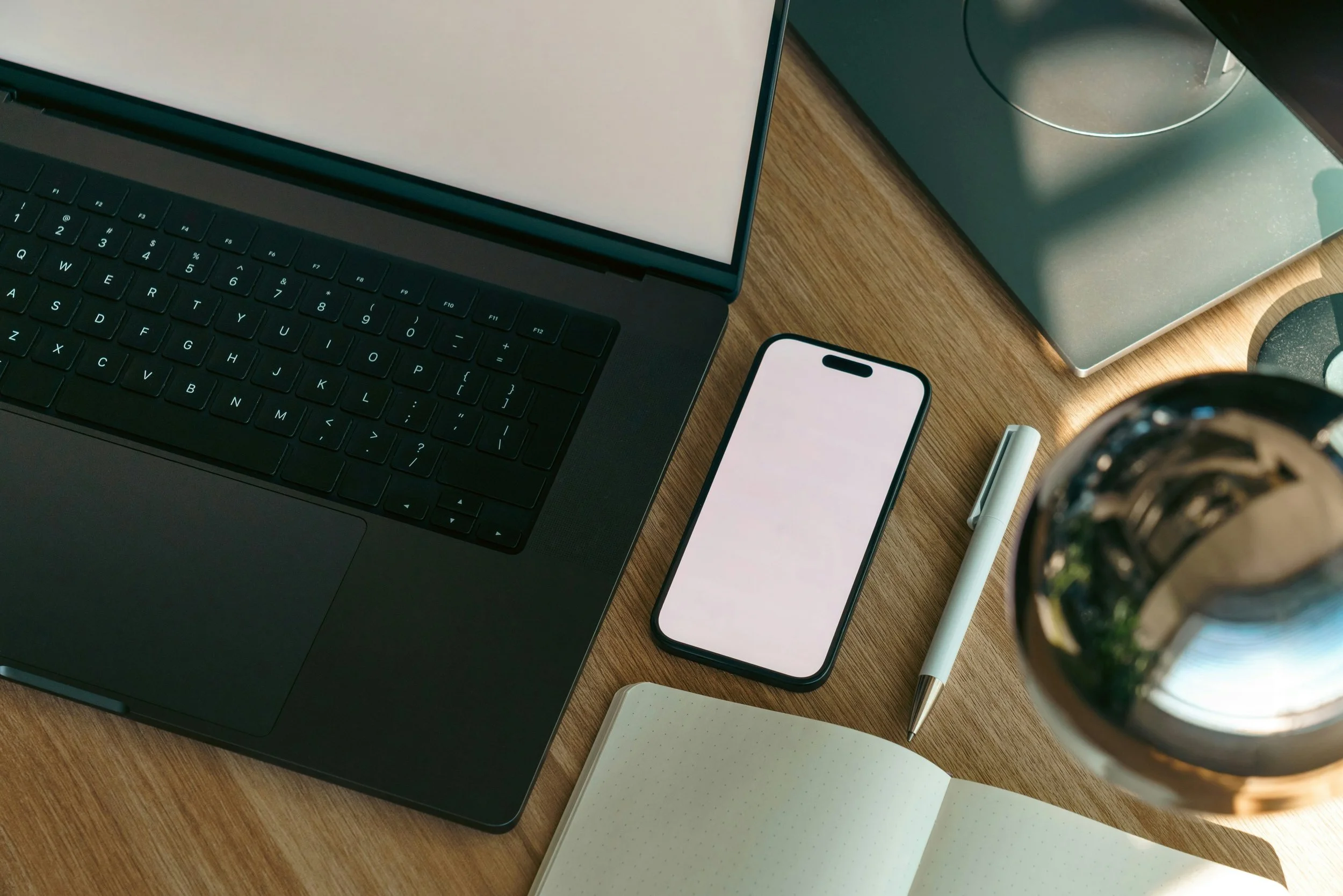 A workspace with a laptop, smartphone, pen, notebook, paper, and a vase with flowers on a wooden desk.