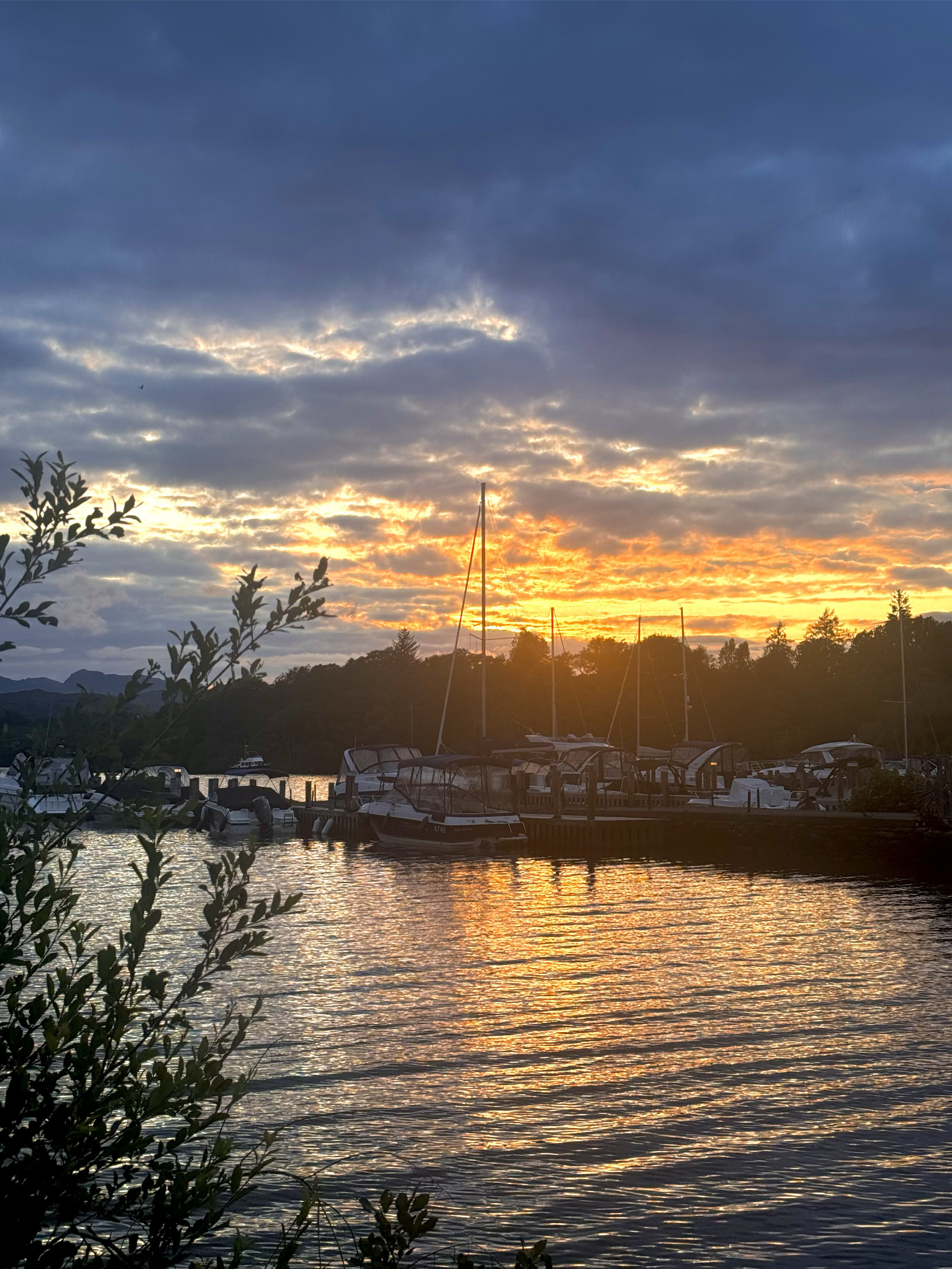 Sunset over a marina with boats docked in calm water, silhouetted trees and hills in the background, and a partly cloudy sky with golden and orange hues.