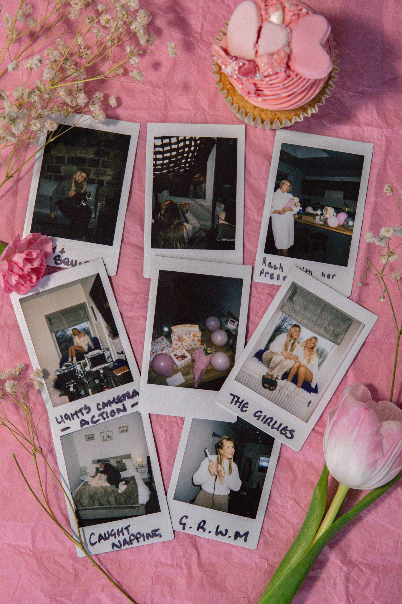 A pink surface with scattered pink and white flowers, several printed Polaroid-style photos, a pink cupcake with heart-shaped toppers, and a pink carnation flower. The photos have handwritten labels such as 'Caught in Happiness,' 'The Gubbles,' and i