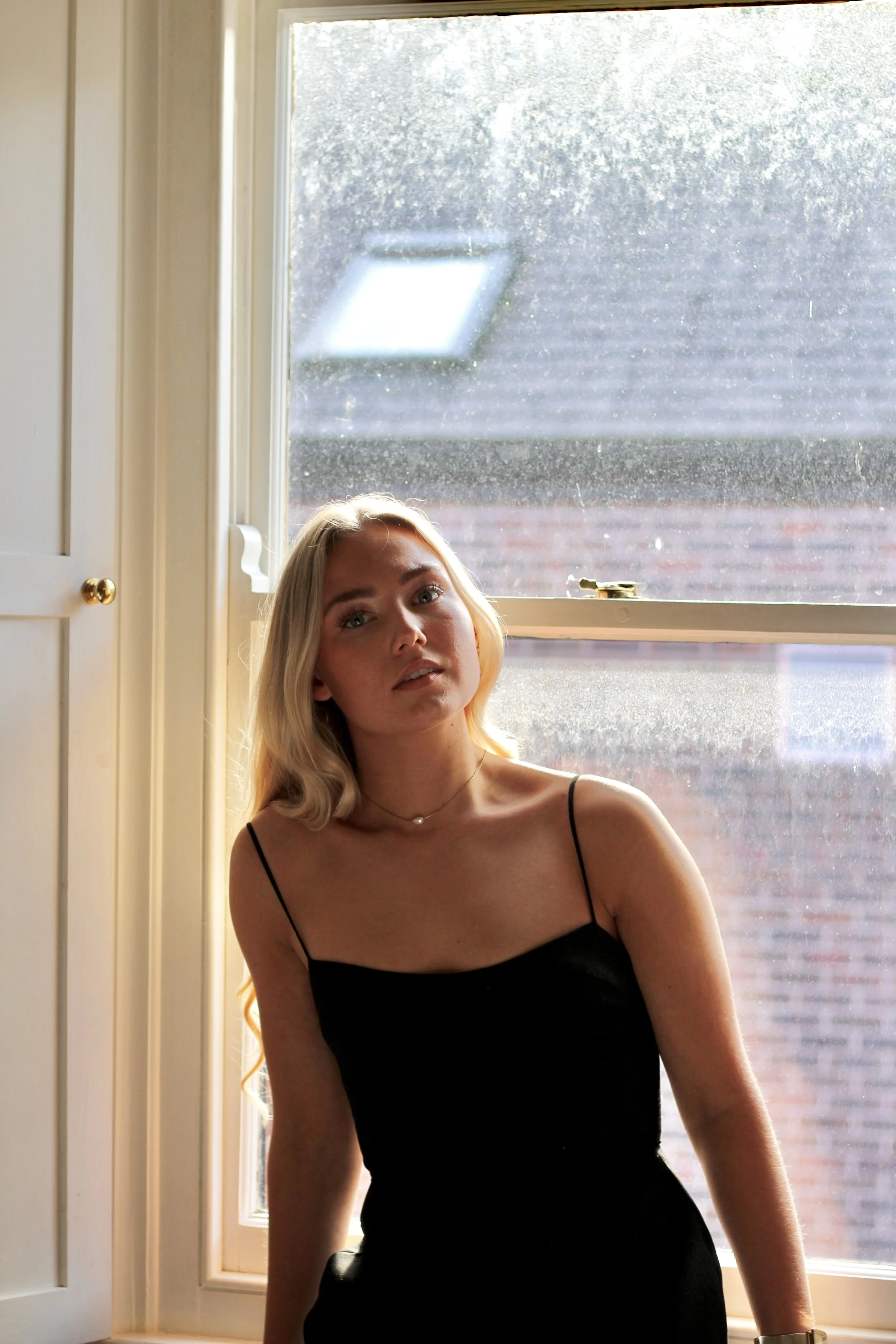 A young woman with long blonde hair and fair skin stands by a window, wearing a black spaghetti strap dress, looking slightly upwards with a neutral expression, with sunlight streaming through the window behind her.