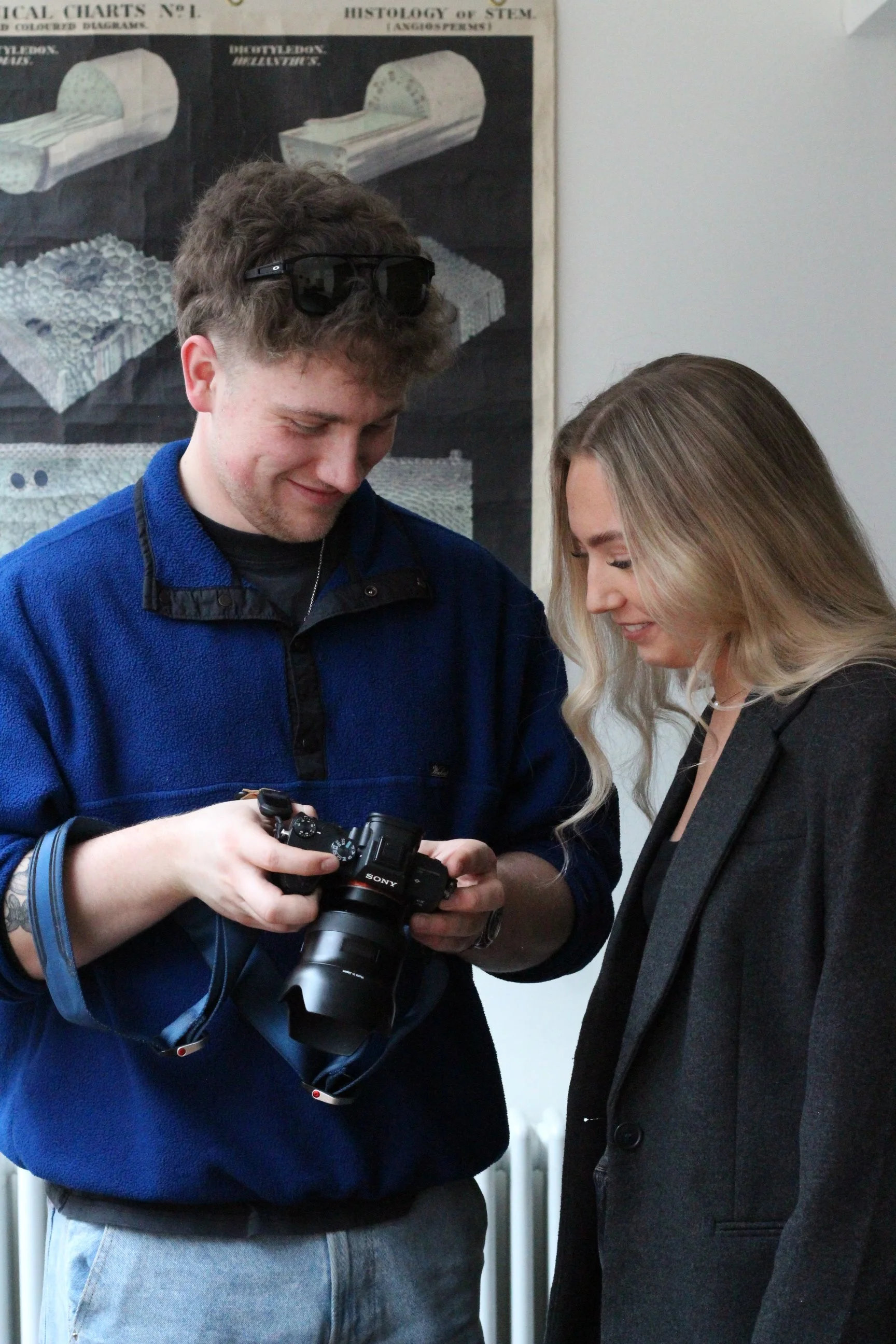A young man with curly hair and sunglasses on his head is showing a woman something on a Sony camera. They are both smiling and standing indoors near a wall with a medical diagram poster.