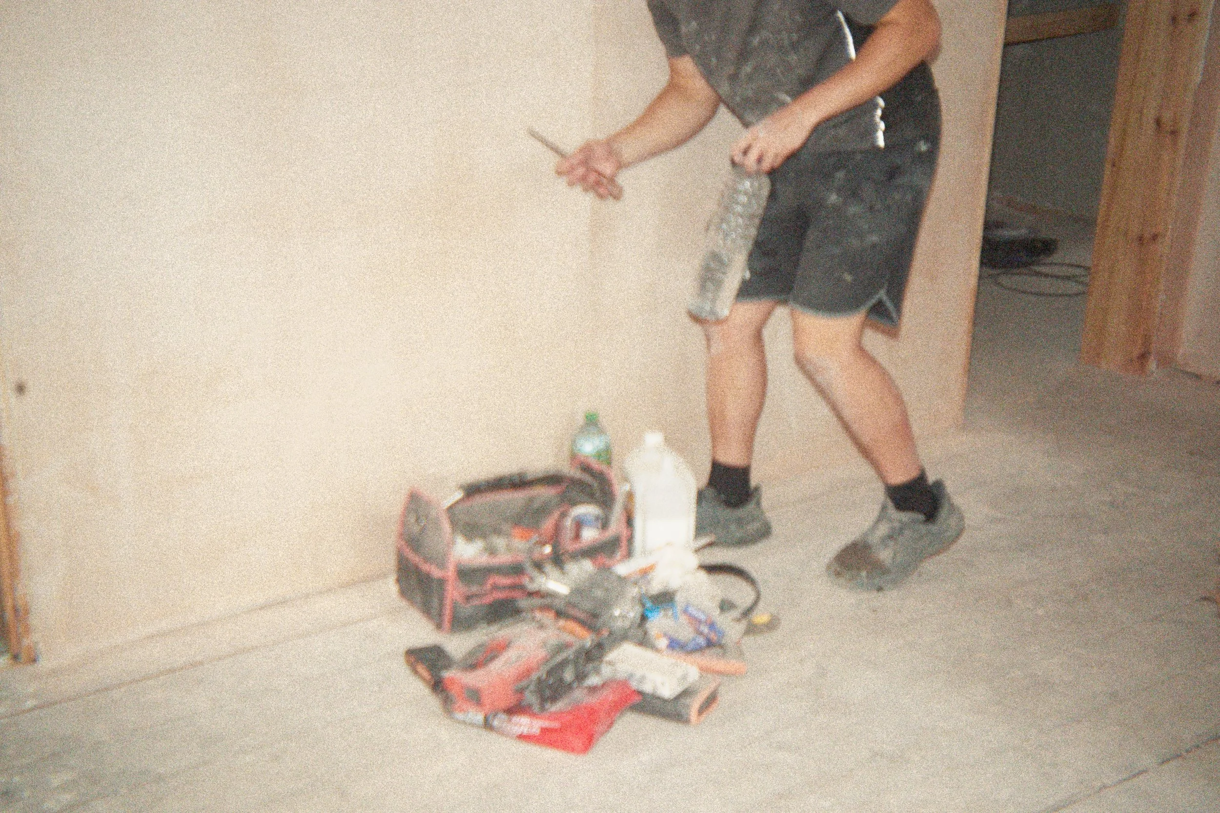 A person in a gray shirt and black shorts is painting a wall with a paint roller. There are various items on the floor including a toolbox, a bottle, and several tools.