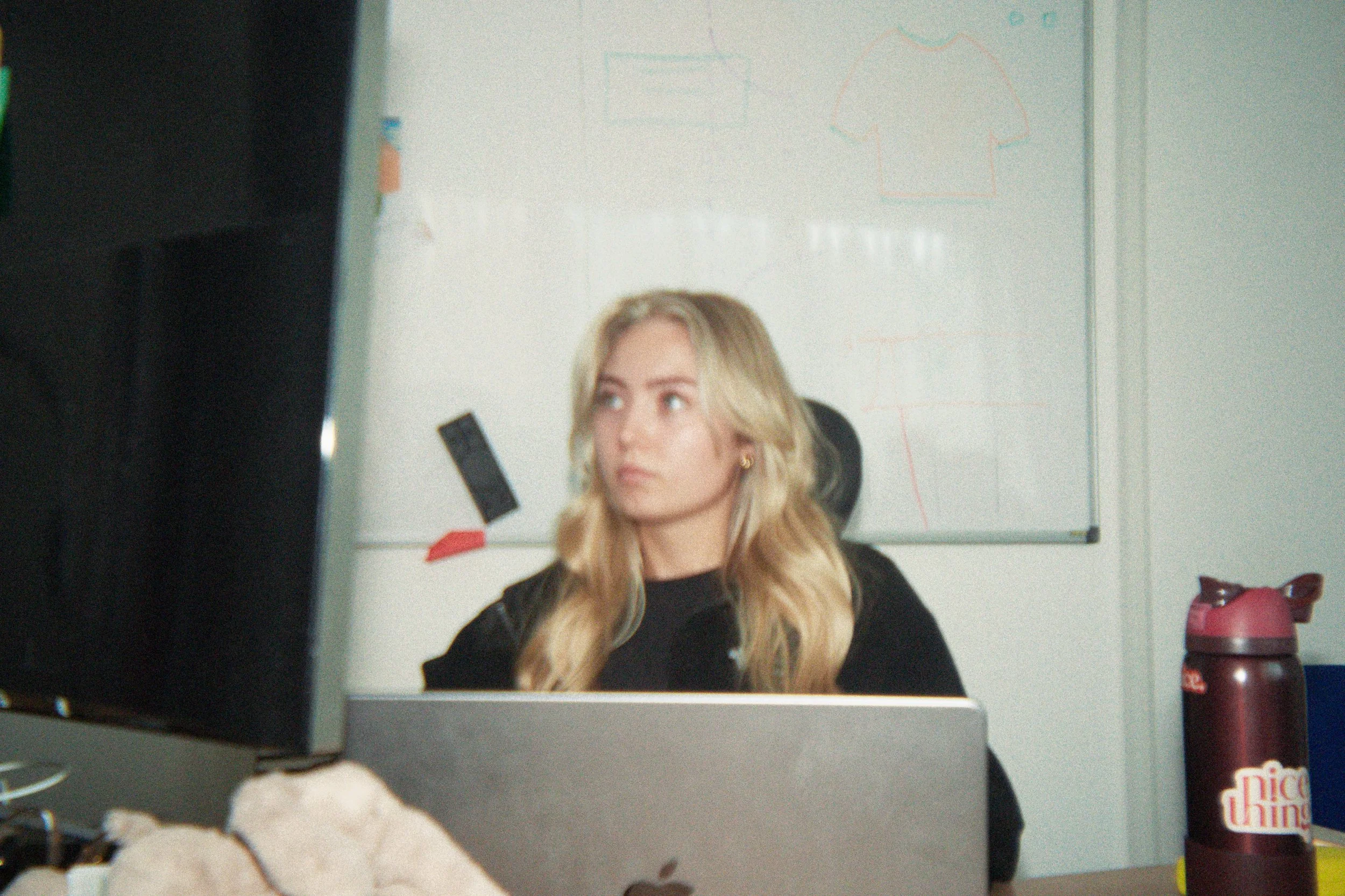 A woman with blonde hair sitting in front of a computer, looking to her left. There is a whiteboard with drawings or notes behind her. In the foreground, there is a partially eaten cookie and a pink water bottle on the desk.
